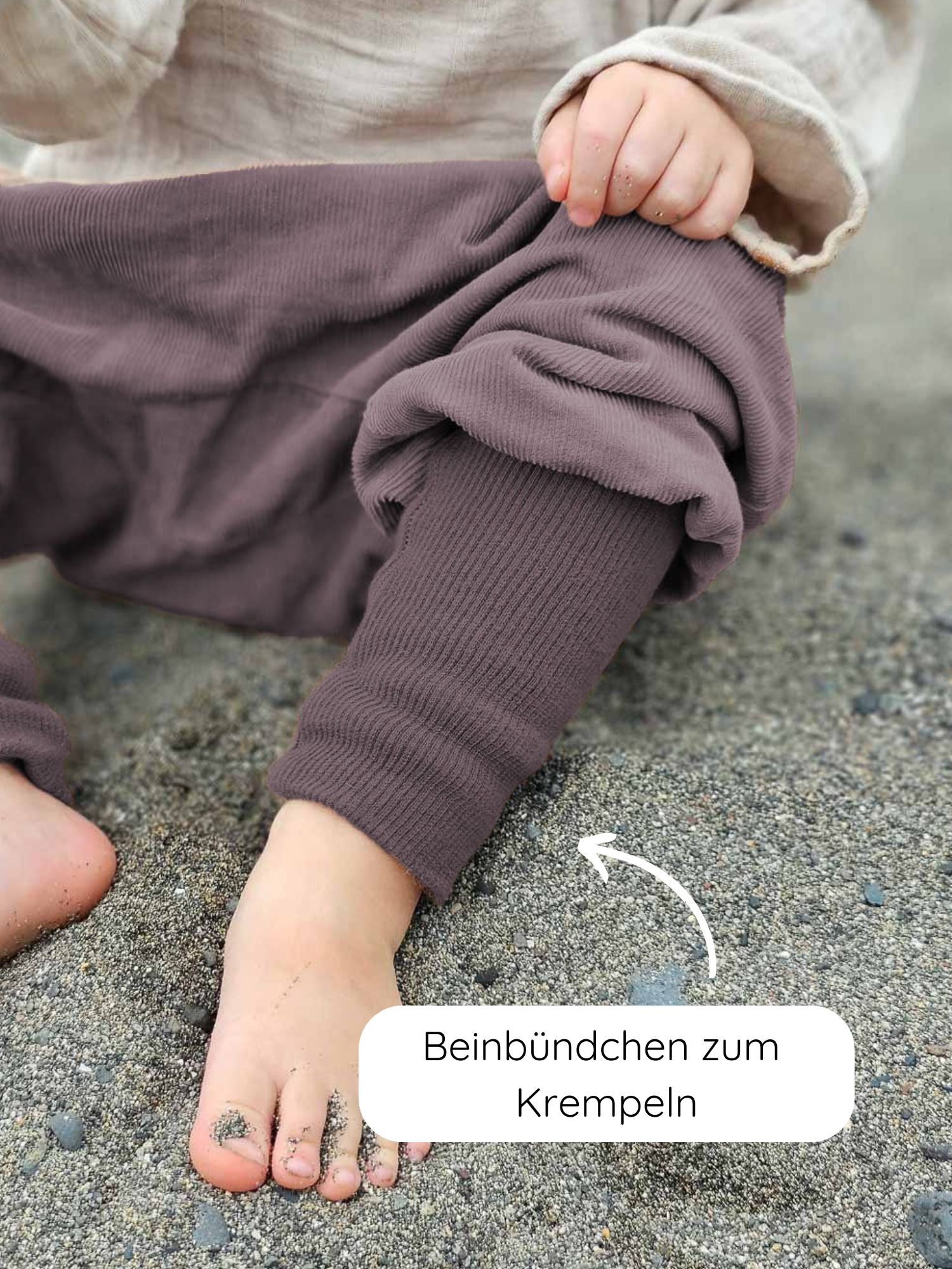 Knickerbocker Feincord - Malve: Nahaufnahme von Kinderfüßen auf sandigem Boden, mit hochgekrempelten, gerippten Beinbündchen. Ein weißer Pfeil zeigt auf die Bündchen mit dem Hinweis Beinbündchen zum Krempeln.