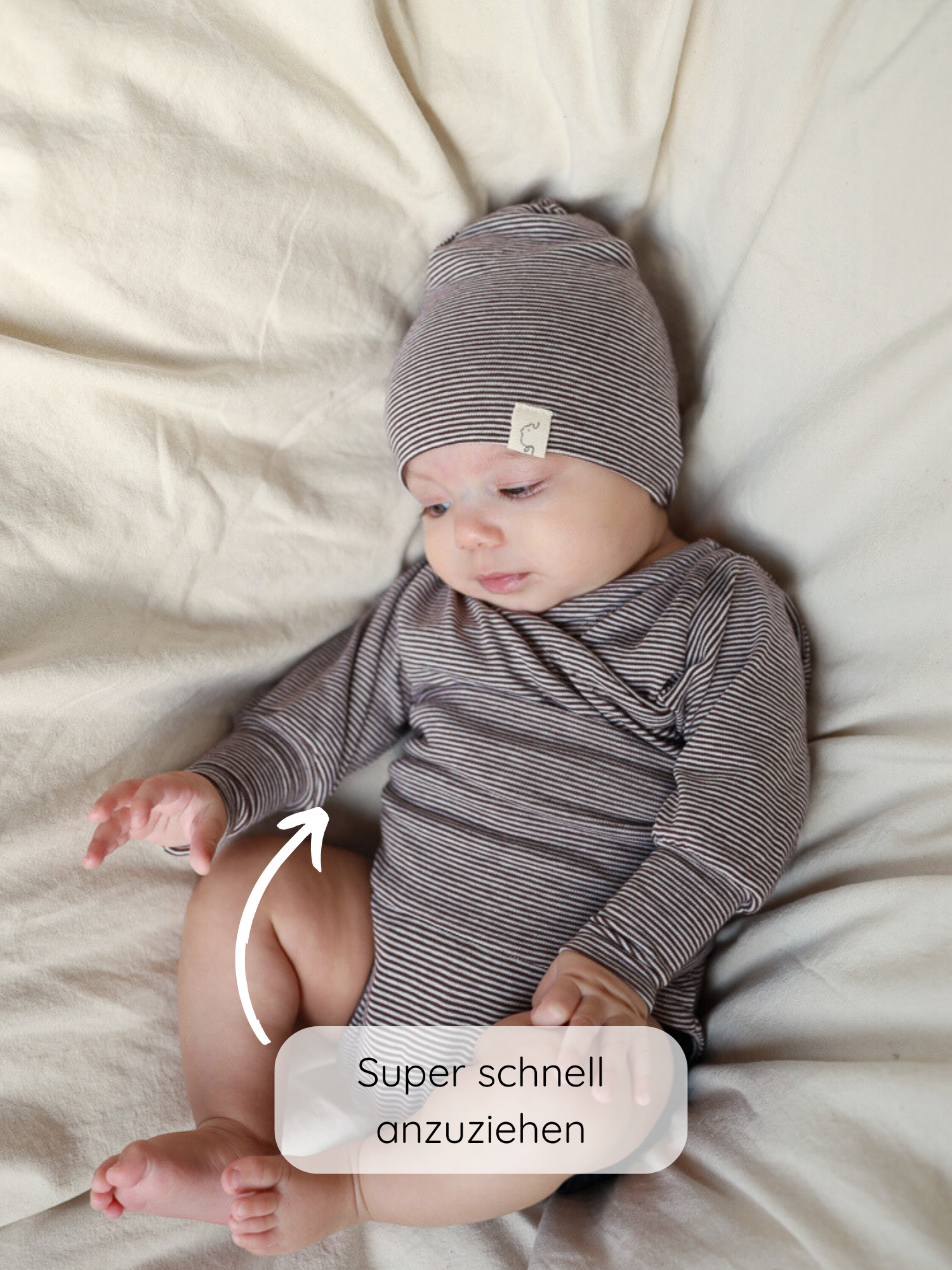Baby in gestreiftem Loopbody Bio-Baumwolle-Ringel Schoko auf cremefarbener Decke, mit Beanie. Weißer Pfeil zeigt auf den Halsausschnitt, Text Super schnell anzuziehen über dem Bild.