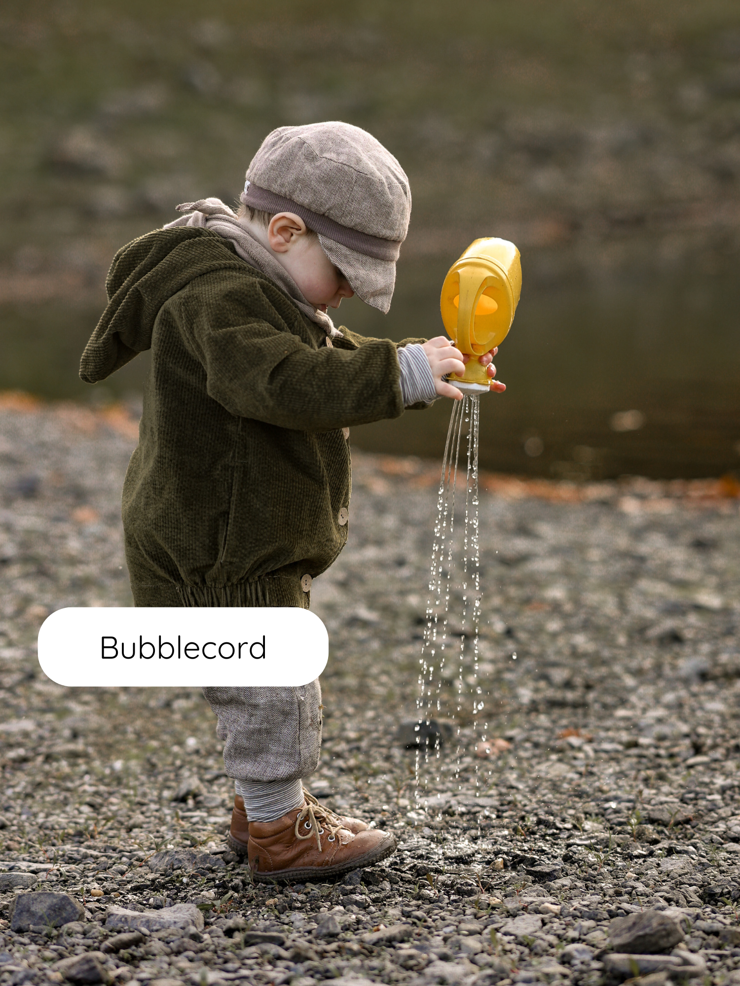 Ein Kleinkind am Flussufer trägt die Wichteljacke Breitcord Stretch - Kiwi mit süßer Wichtelkapuze, kombiniert mit Ringel-Armbündchen, während es mit einer gelben Gießkanne Wasser auf die Steine gießt.