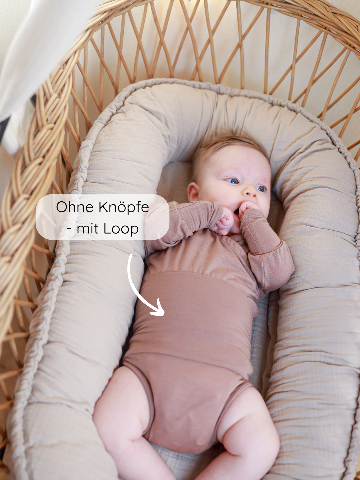 Kleinkind in einem Loopbody Kaschmir-Mix - Puder, ohne Knöpfe, mit Loop am Bauch für einfaches Anziehen. Liegt auf gepolsterter Matratze im geflochtenen Wicker-Bassinet.