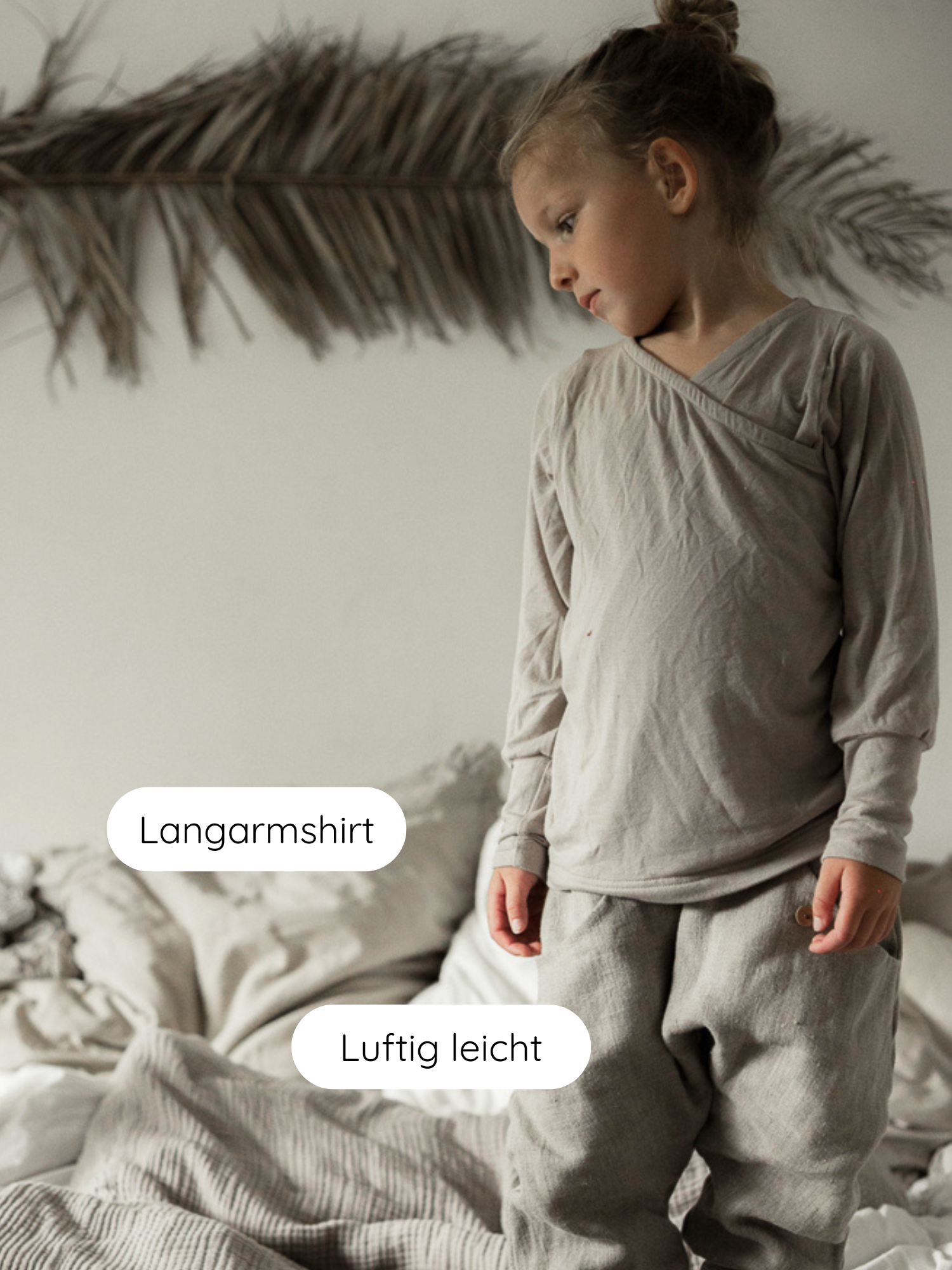 Kleinkind auf Bett trägt Wickelshirt aus Kaschmir-Mix - Sand mit luftigen, weiten Hosen. Umgeben von natürlicher, minimalistischer Einrichtung mit Bettwäsche und getrockneter Palmblätter-Deko an der Wand.