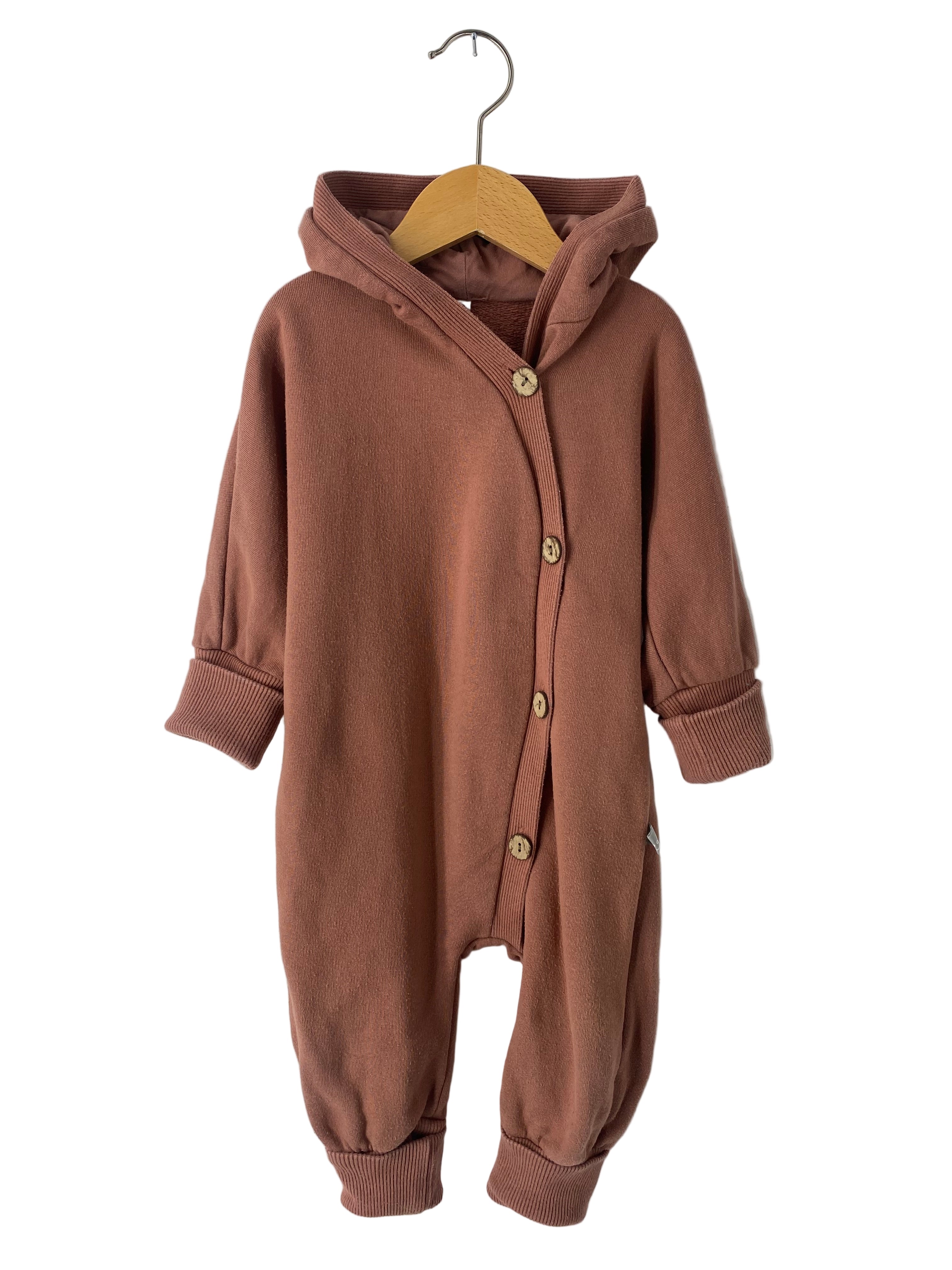 Kuschelanzug Bio-Sweat Zimt Gr 6M: Einteiler mit Kapuze, langen Ärmeln, gerippten Bündchen und Holzoptik-Knöpfen, präsentiert auf einem Holzkleiderbügel vor neutralem Hintergrund. Zustand: gut.