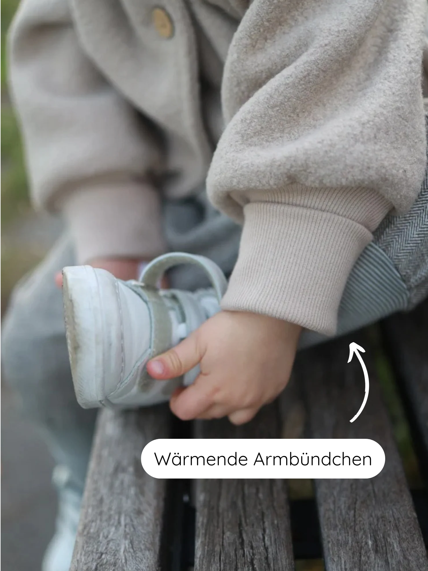 Kleinkind auf Holzbank hält hellen Sneaker, trägt die Wichteljacke Wollwalk - Sand mit wärmenden Armbündchen aus Bio-Baumwolle. Pfeil zeigt auf gerippten Ärmelbund, hervorgehoben als Wärmende Armbündchen.