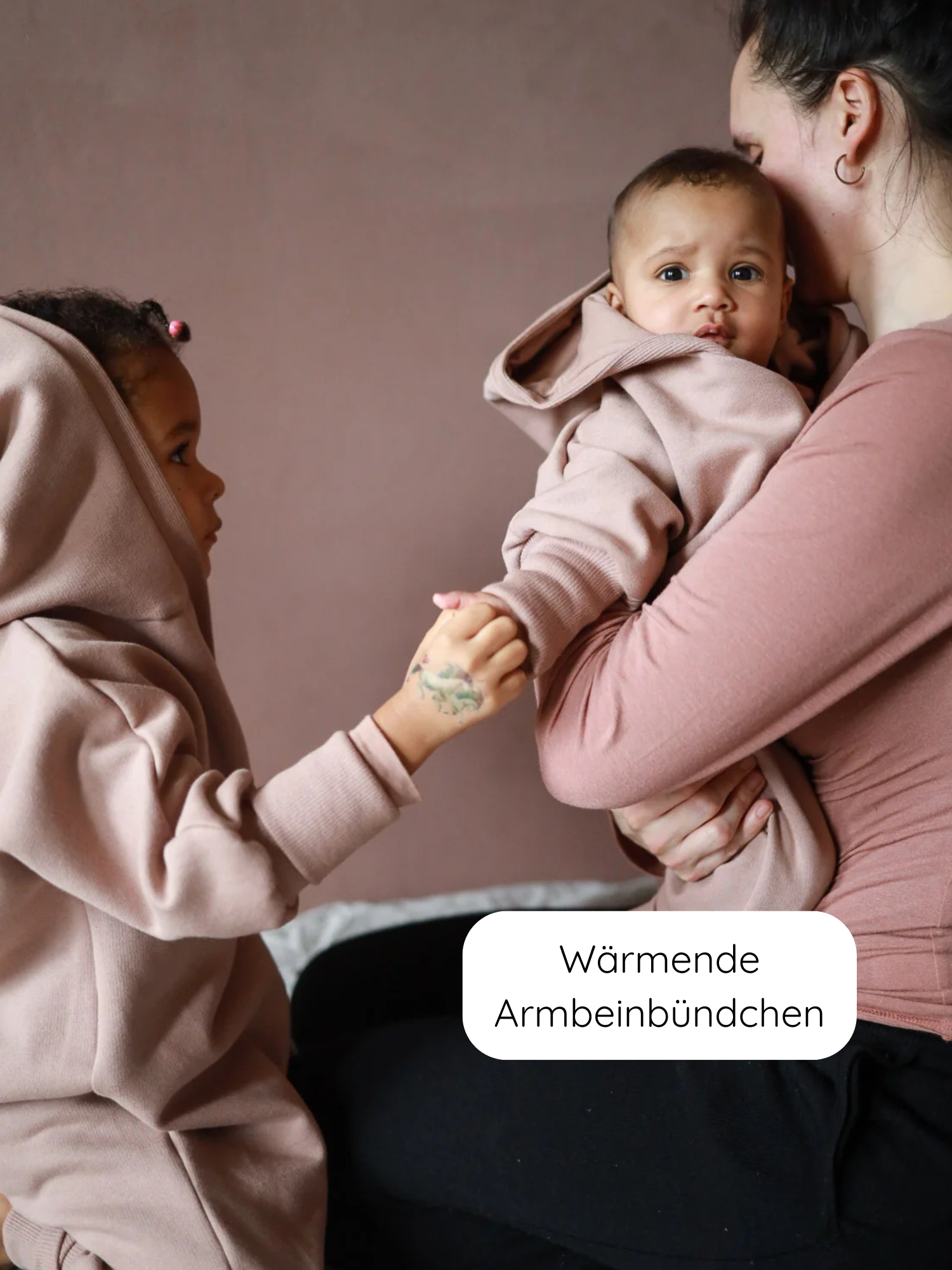 Frau hält Baby im Kuschelanzug Bio-Sweat - Puder mit Kapuze, während älteres Kind die Hand des Babys hält. Wärmende Arm- und Beinbündchen betonen den Komfort und die Qualität des Anzugs.