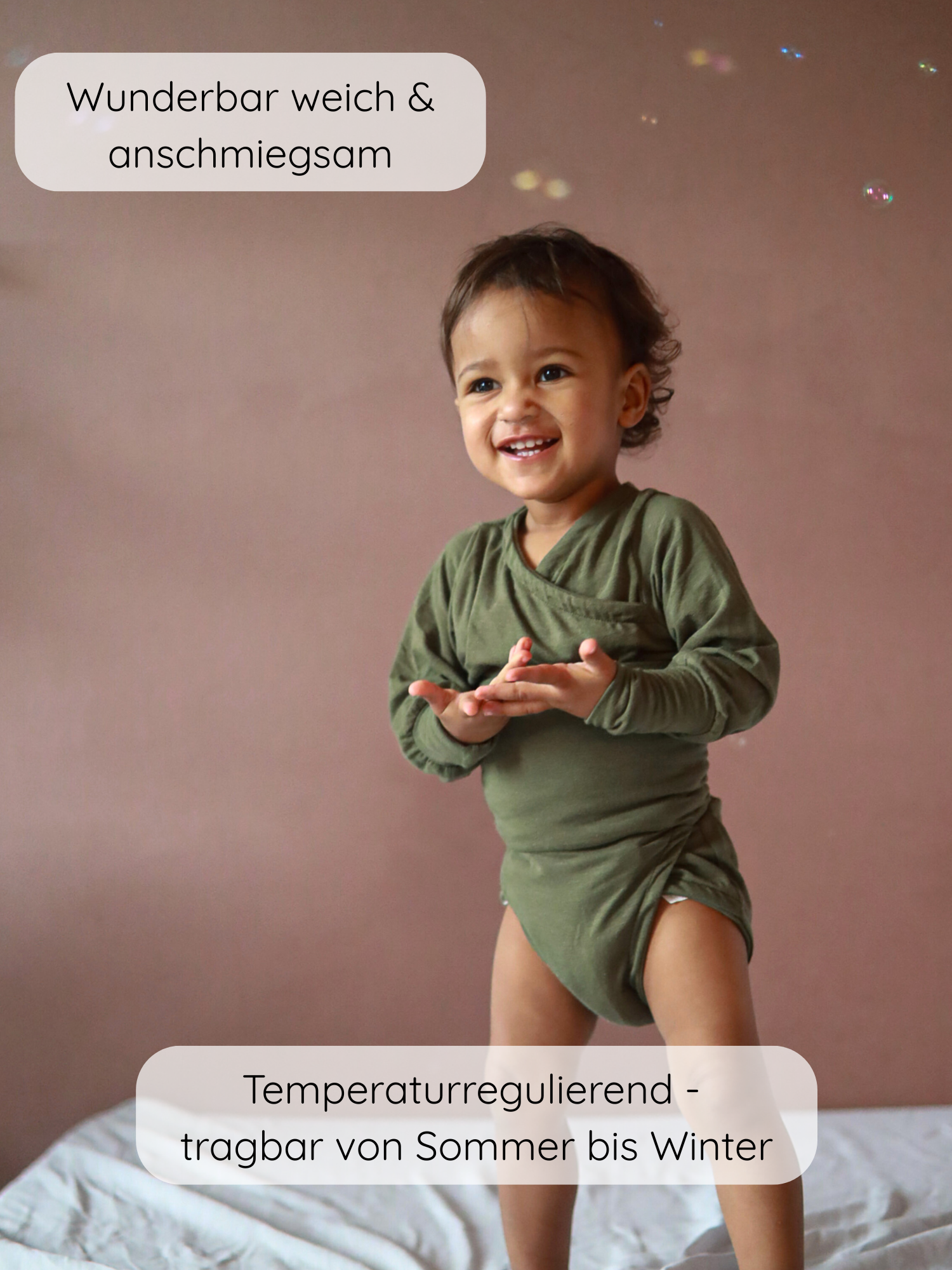 Kleinkind in einem Loopbody Kaschmir-Mix - Pistazie, lächelnd, auf einem Bett stehend, mit schwebenden Seifenblasen. Der Body ist langärmelig und ohne Knöpfe, aus weichem Kaschmir-Mix.