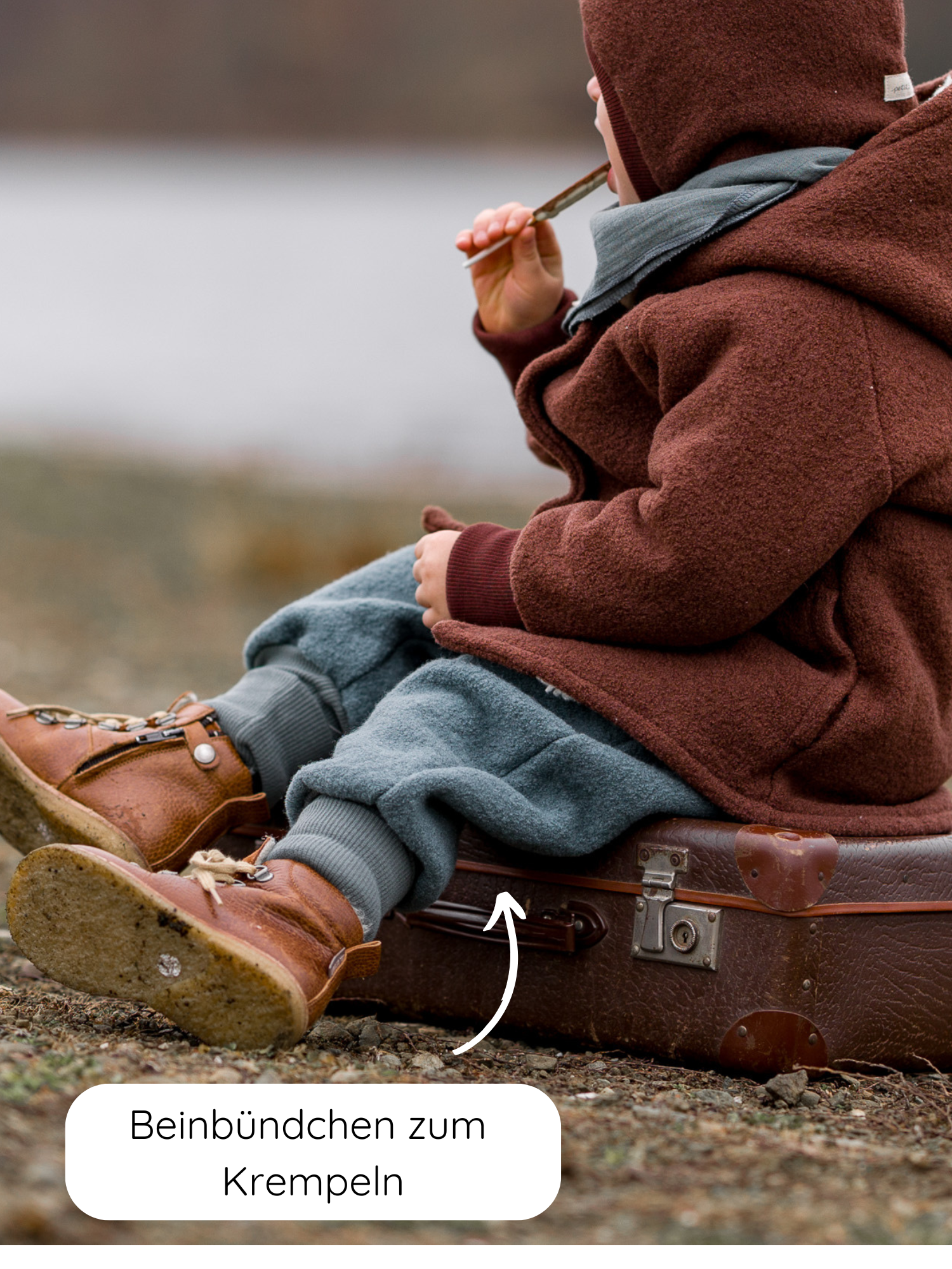 Ein kleines Kind sitzt auf einem braunen Koffer und trägt die Knickerbocker Wollwalk - Taube mit aufrollbaren Beinbündchen, kombiniert mit tanfarbenen Lederstiefeln. Ein Pfeil zeigt auf die krempelbaren Bündchen.