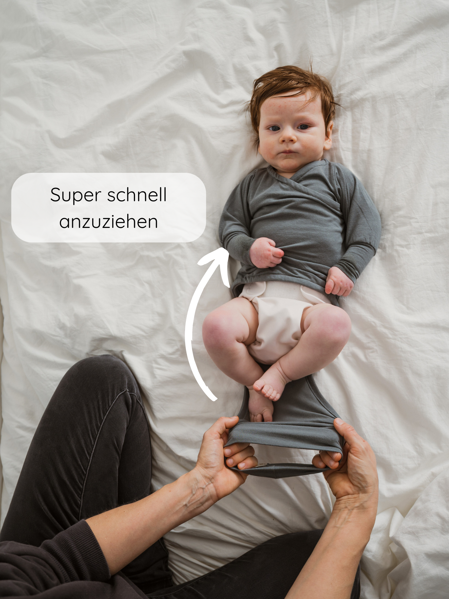 Ein Baby trägt einen Loopbody Kaschmir-Mix - Taube. Eine erwachsene Hand zieht den weichen, grauen Langarmbody über die Beine des Babys. Ein Pfeil zeigt, wie leicht er anzuziehen ist.