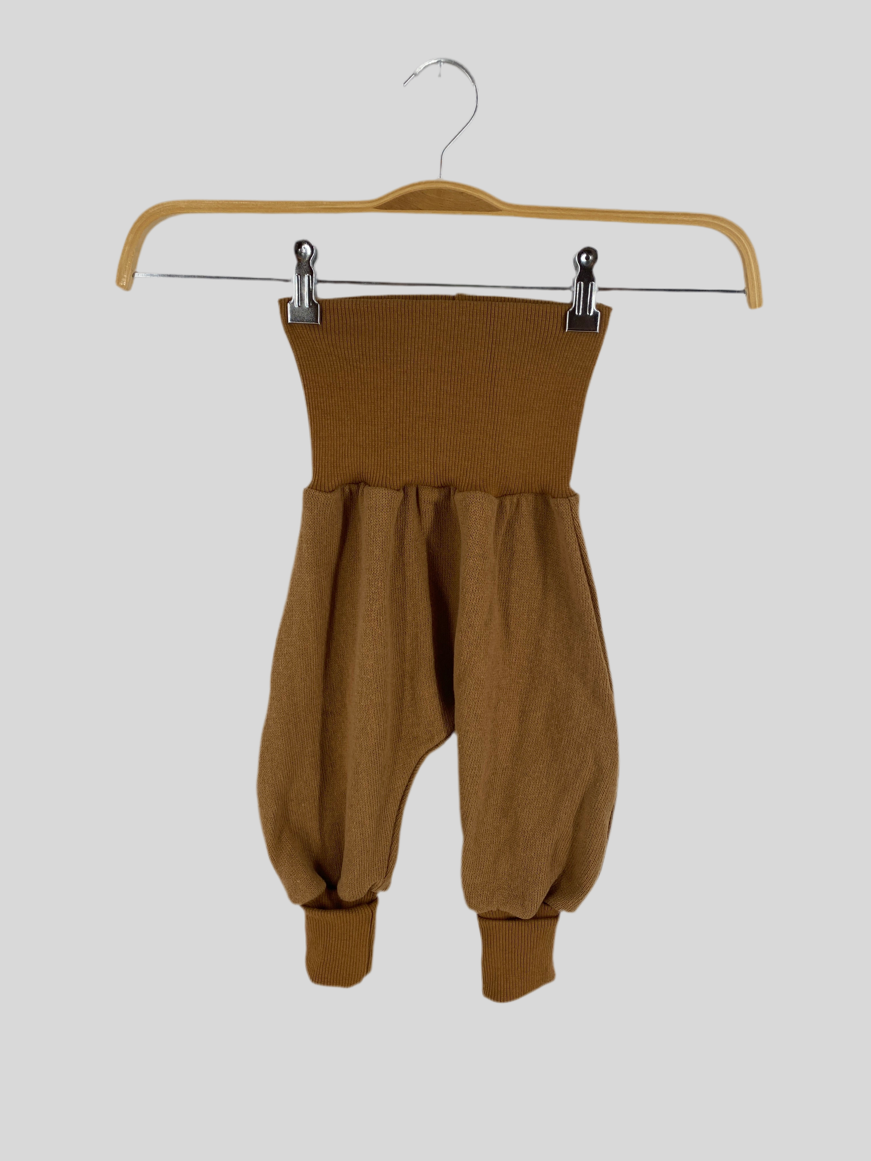 Babyknicker Strick Sahara mit Rippbündchen Gr 1Y: Locker geschnittene Haremshose für Babys aus geripptem Strickstoff, mit breitem, dehnbarem Bund und schmalen Bündchen, präsentiert an einem Holzkleiderbügel vor grauem Hintergrund.