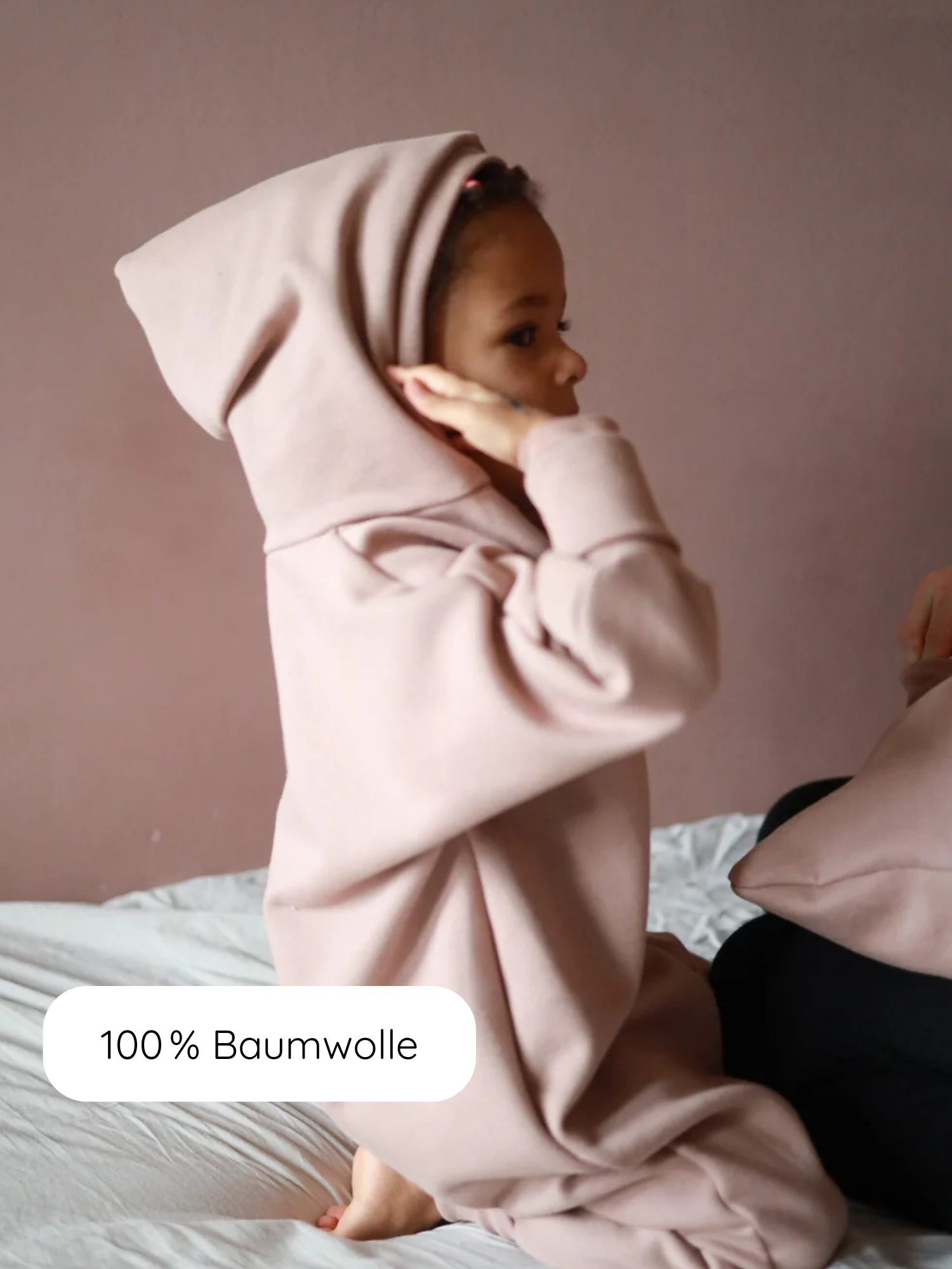 Ein Kind trägt den Kuschelanzug Bio-Sweat - Puder, einen weichen, übergroßen Kapuzenpullover aus 100 % Baumwolle, kniet auf einem Bett und zeigt das bequeme, gemütliche Design in Profilansicht.