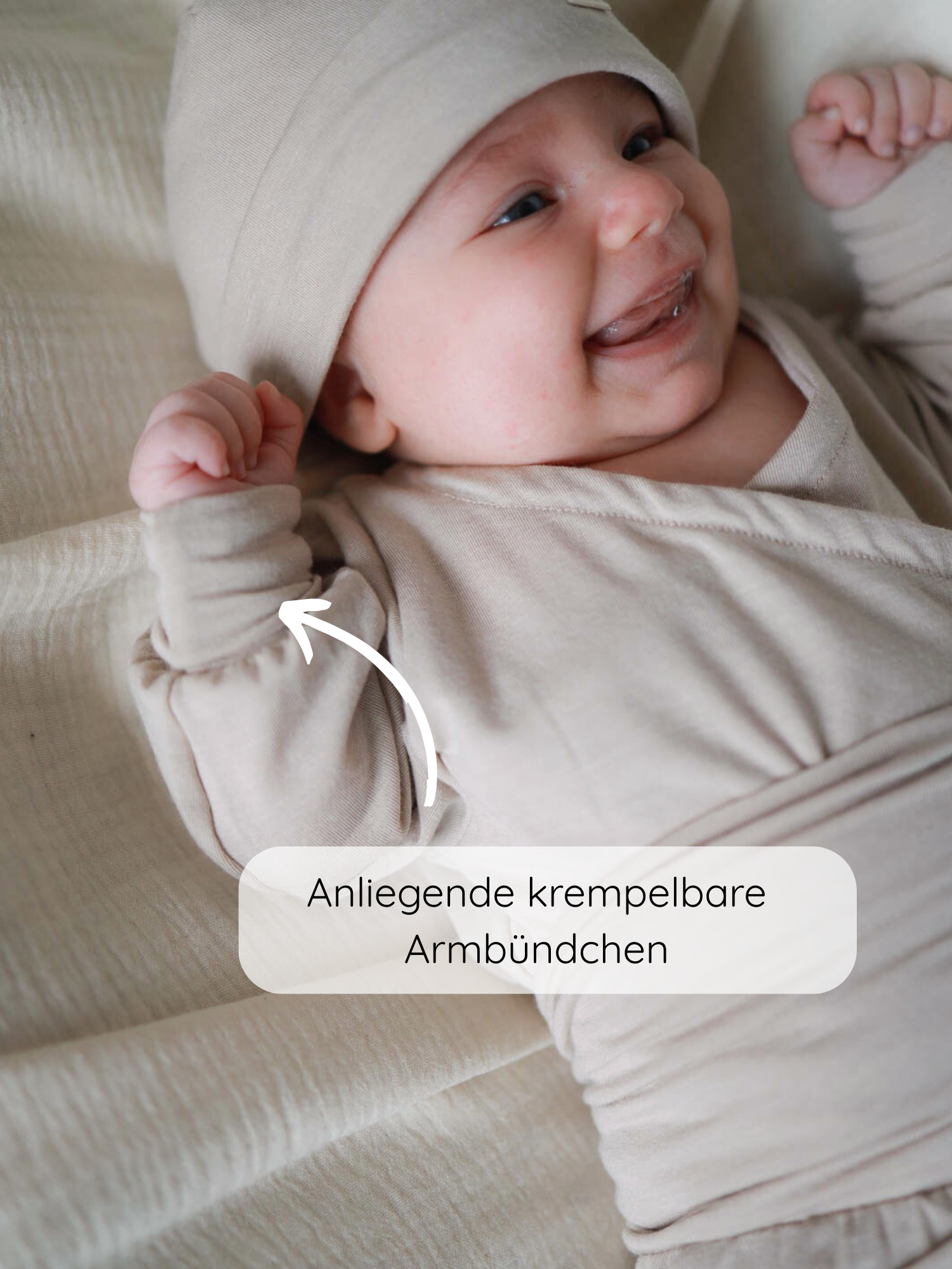 Ein lächelndes Baby trägt den Loopbody Kaschmir-Mix - Sand, mit anliegenden krempelbaren Armbündchen, auf einem neutralen Stoff liegend. Ein Pfeil zeigt auf die besondere Ärmelkonstruktion.