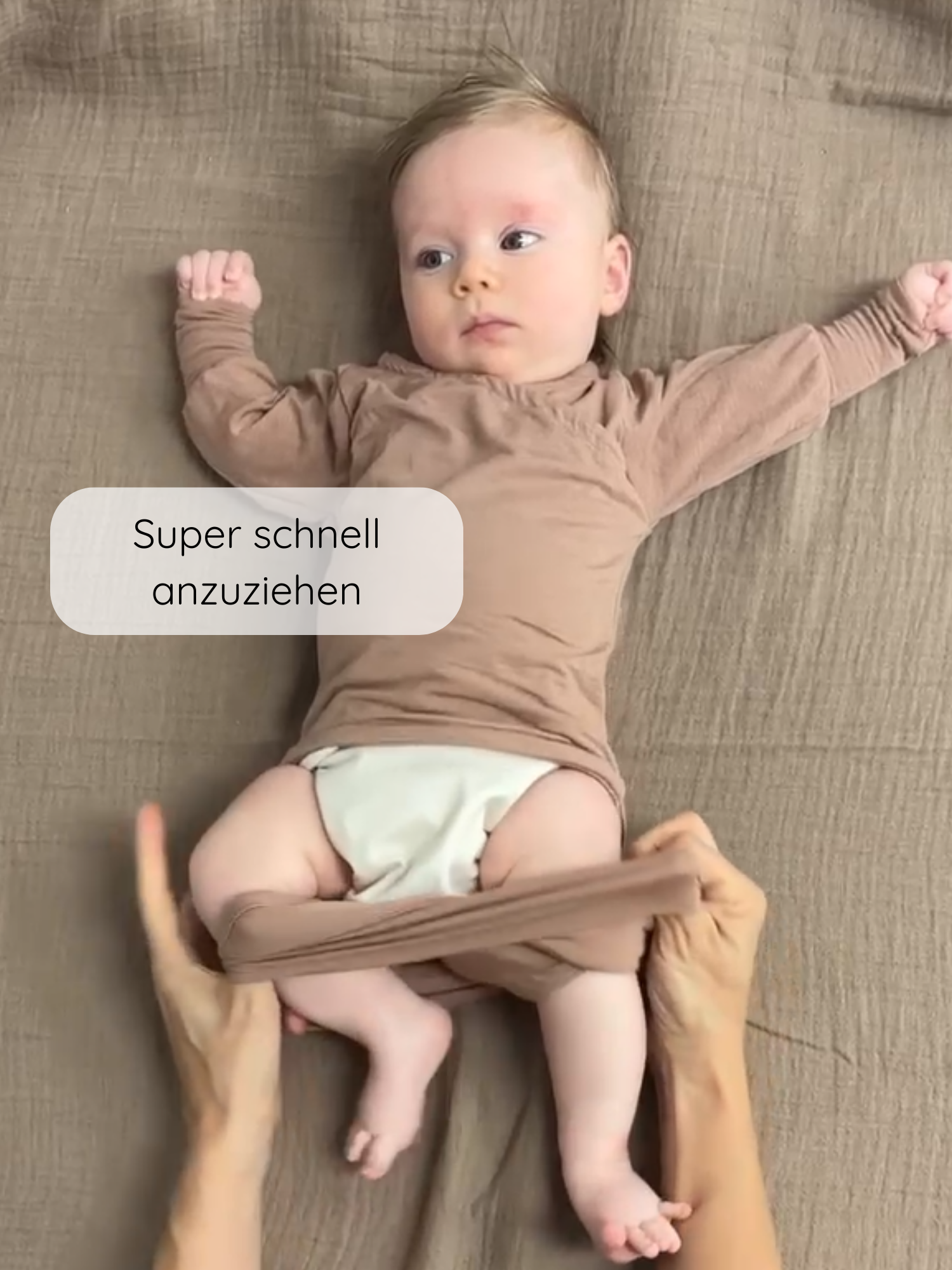 Baby in einem Loopbody Kaschmir-Mix - Puder, der super schnell anzuziehen ist. Zwei Hände ziehen das weiche, langärmlige Kleidungsstück über die Beine des Babys für einfachen Komfort.