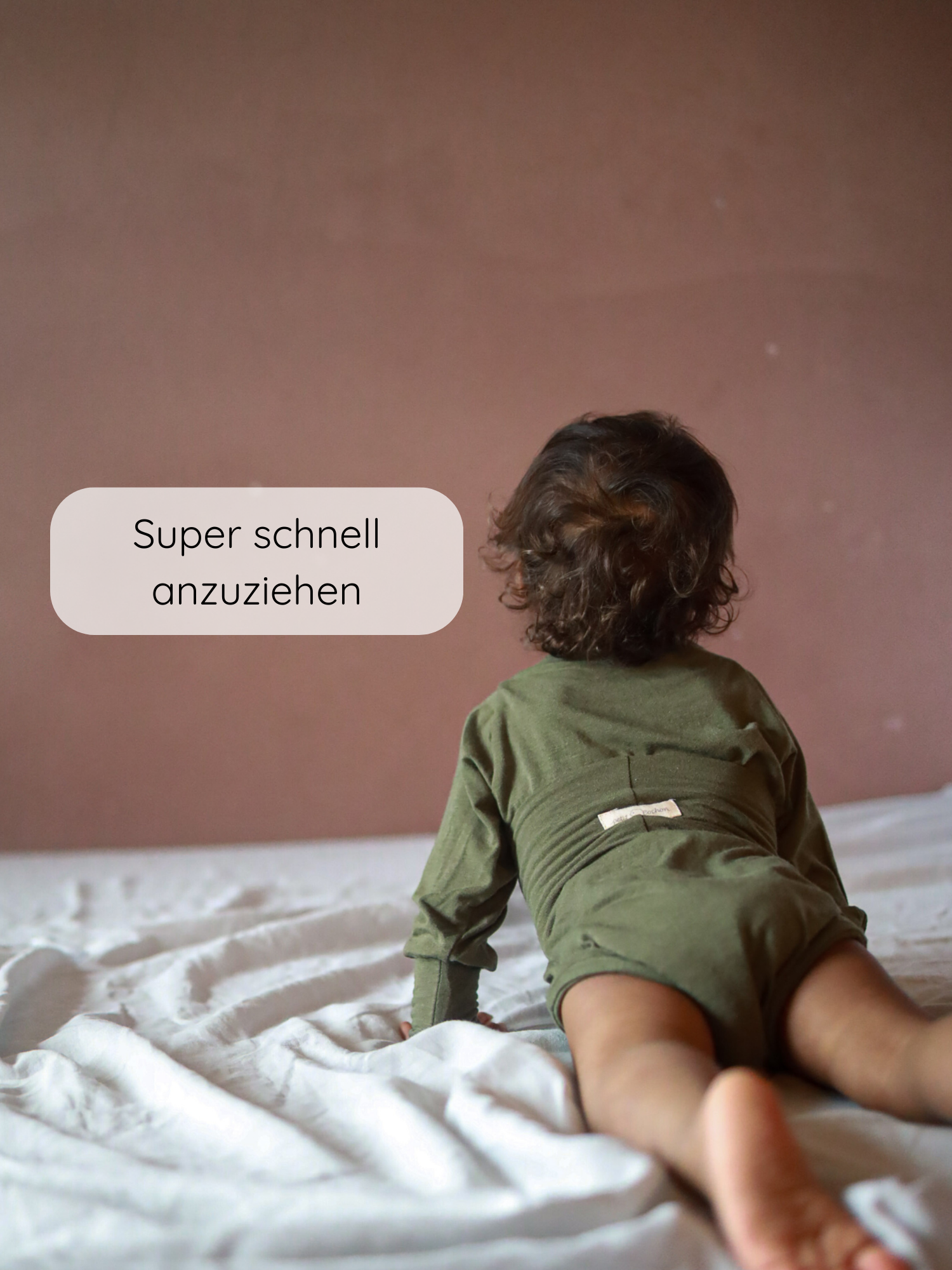 Kleinkind krabbelt auf Bett, trägt Loopbody Kaschmir-Mix - Pistazie. Textblase „Super schnell anzuziehen“ zeigt die einfache Handhabung des weichen, wachsenden Einteilers ohne Knöpfe.