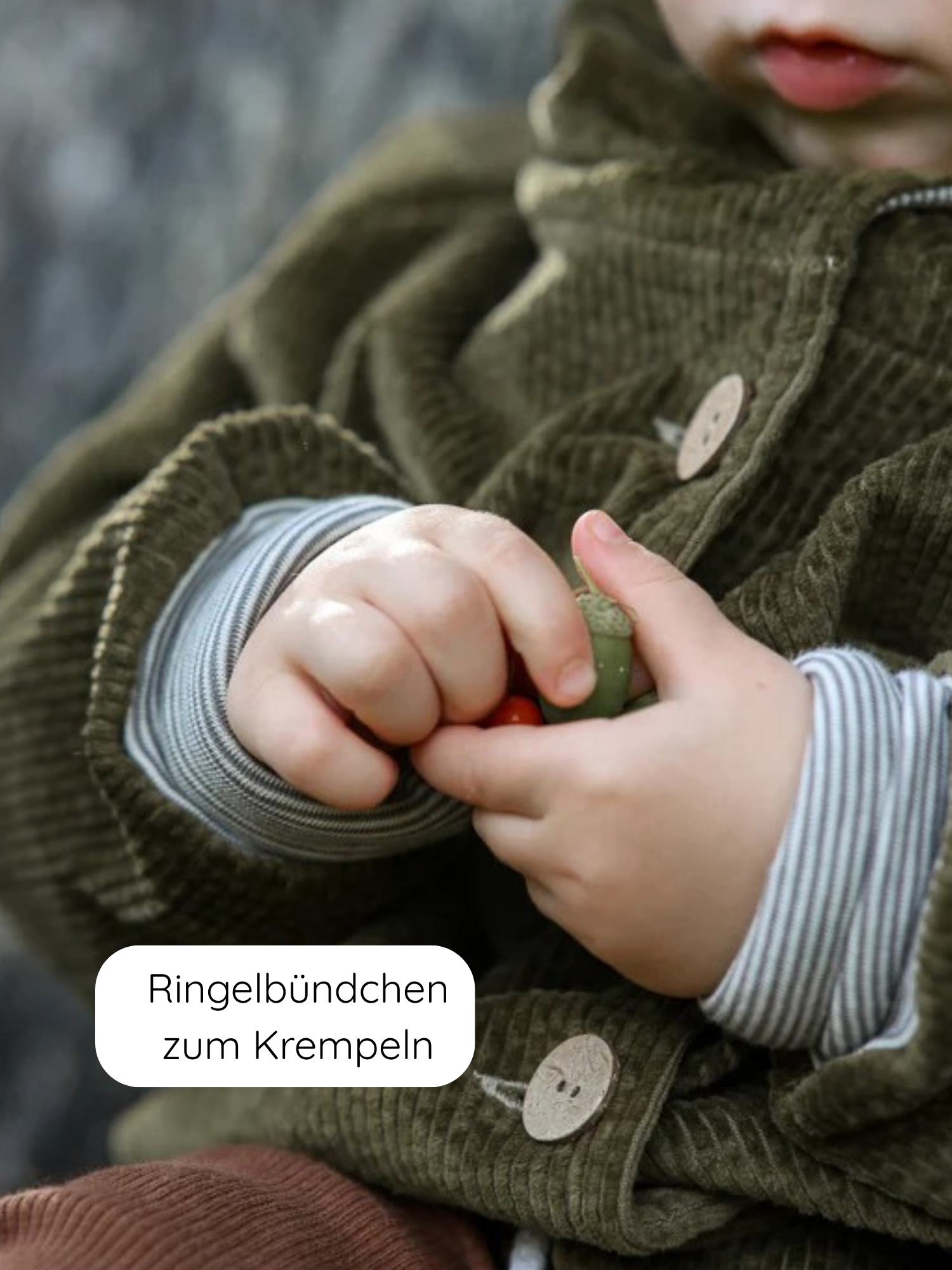 Kleinkind in der Wichteljacke Breitcord Stretch - Kiwi mit großen Holzknöpfen und Ringel-Armbündchen, hält eine Eichel und eine kleine rote Beere.