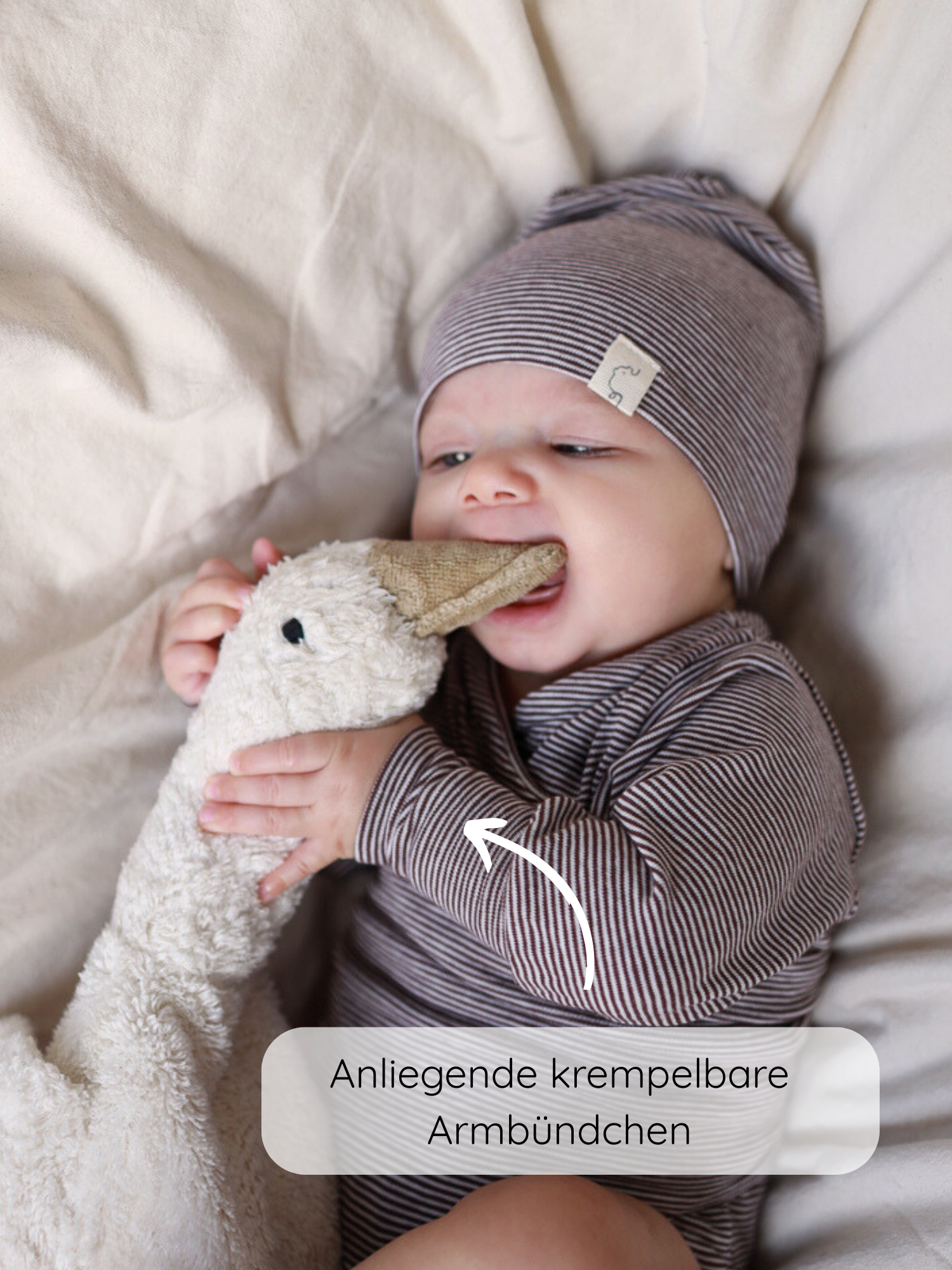 Ein Baby trägt den Loopbody Bio-Baumwolle-Ringel Schoko mit passenden Mütze und spielt mit einem Plüschgans. Ein Pfeil zeigt auf die anliegenden, krempelbaren Armbündchen des Langarmbodys.