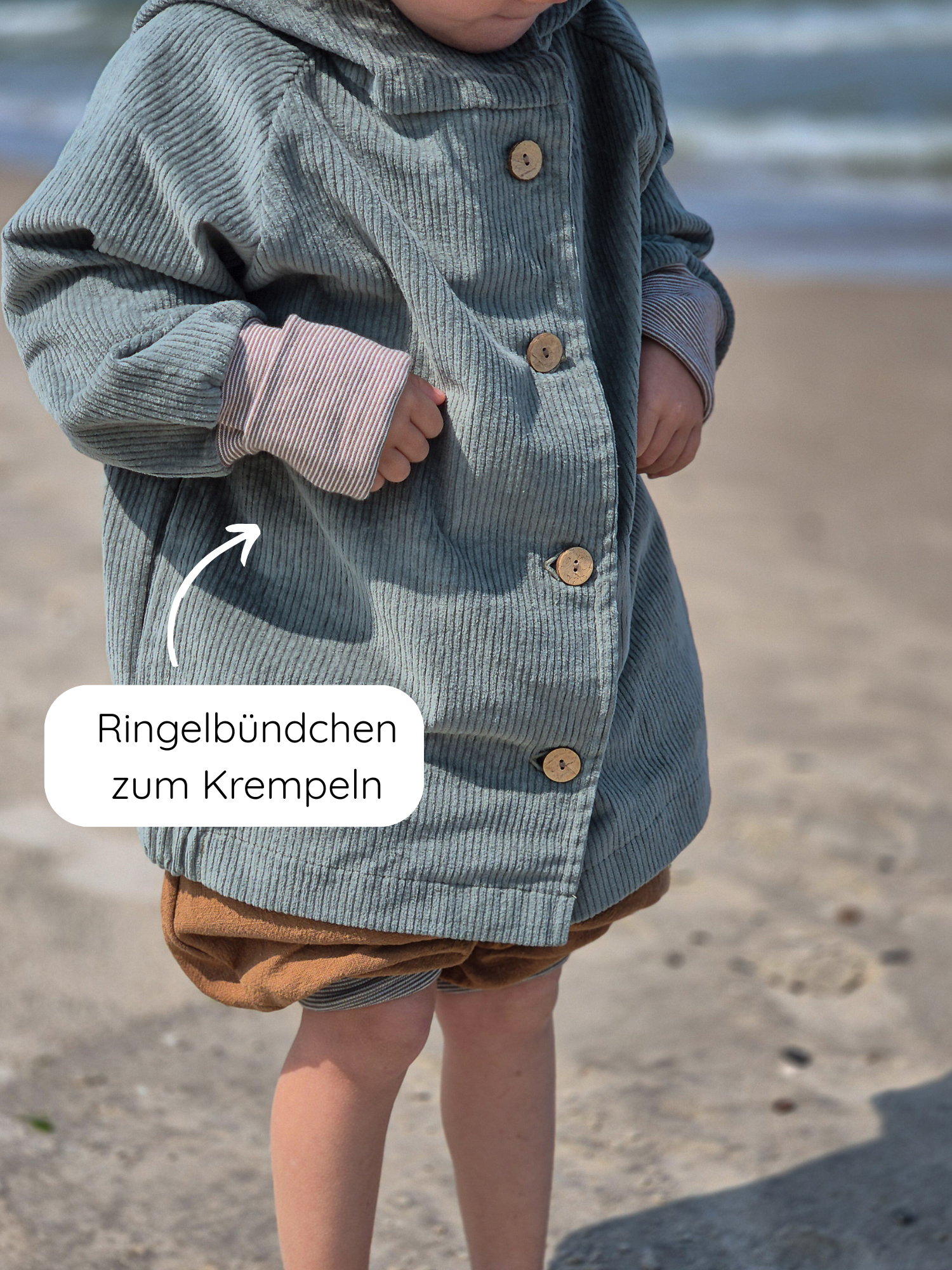 Ein Kind trägt die Wichteljacke Breitcord Stretch- Aqua am Strand. Die Jacke hat Holzknöpfe, krempelbare Ringel-Armbündchen und eine süße Wichtelkapuze, kombiniert mit braunen Shorts.