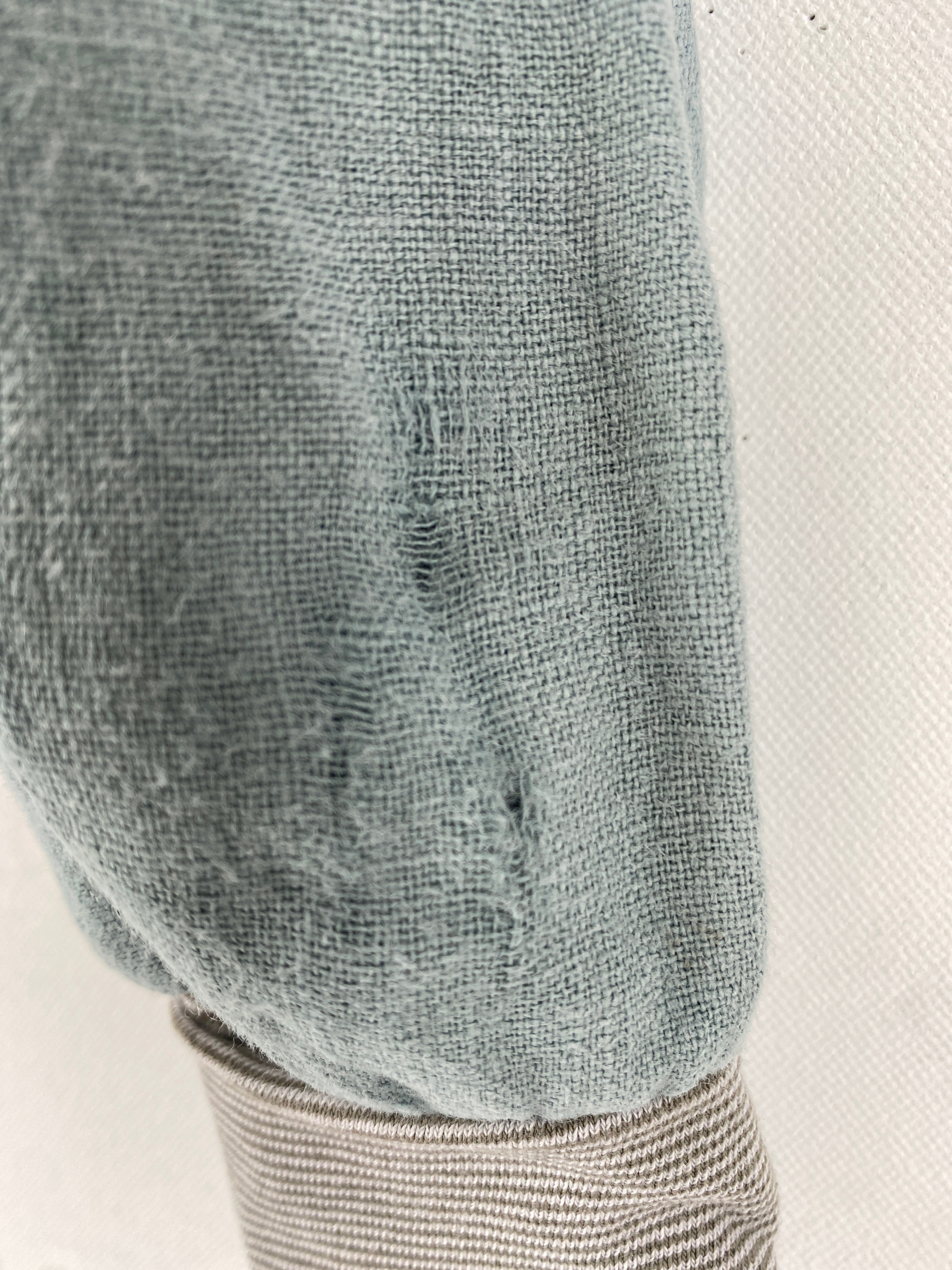 Nahaufnahme eines Knickerbocker Leinen hellblau Gr 6M Ärmels über einem gestreiften Hemd. Der gestrickte Stoff zeigt Webstruktur mit leichten Abnutzungsspuren und dünneren Stellen, Zustand: gut.