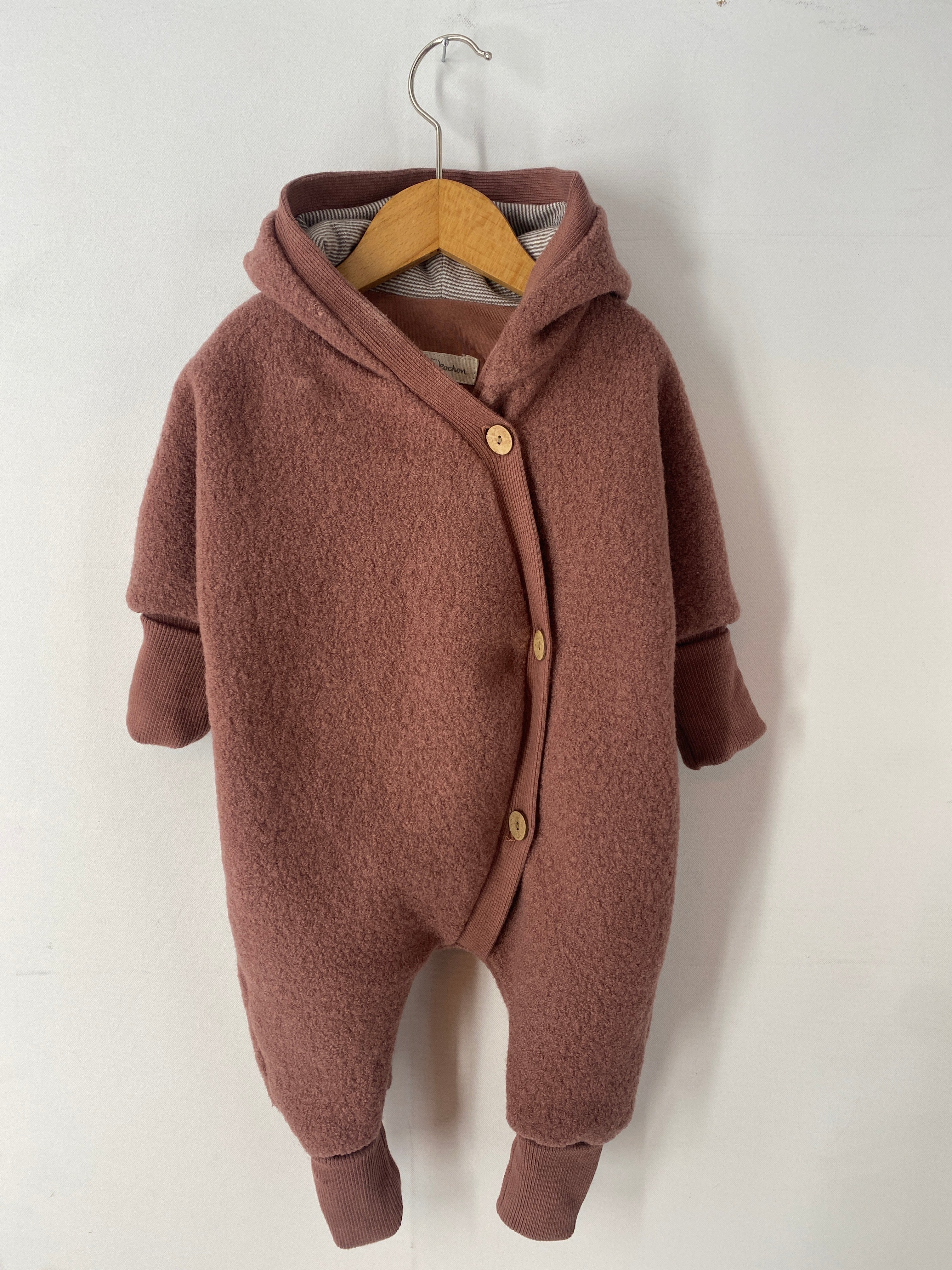 Kuschelanzug Wollwalk Zimt Gr 3M auf einem Kleiderbügel, zeigt sanfte Stoffstruktur und dezente Flecken vorne. Ideal für Babys, bietet es Komfort und Wärme in einem stilvollen Design.