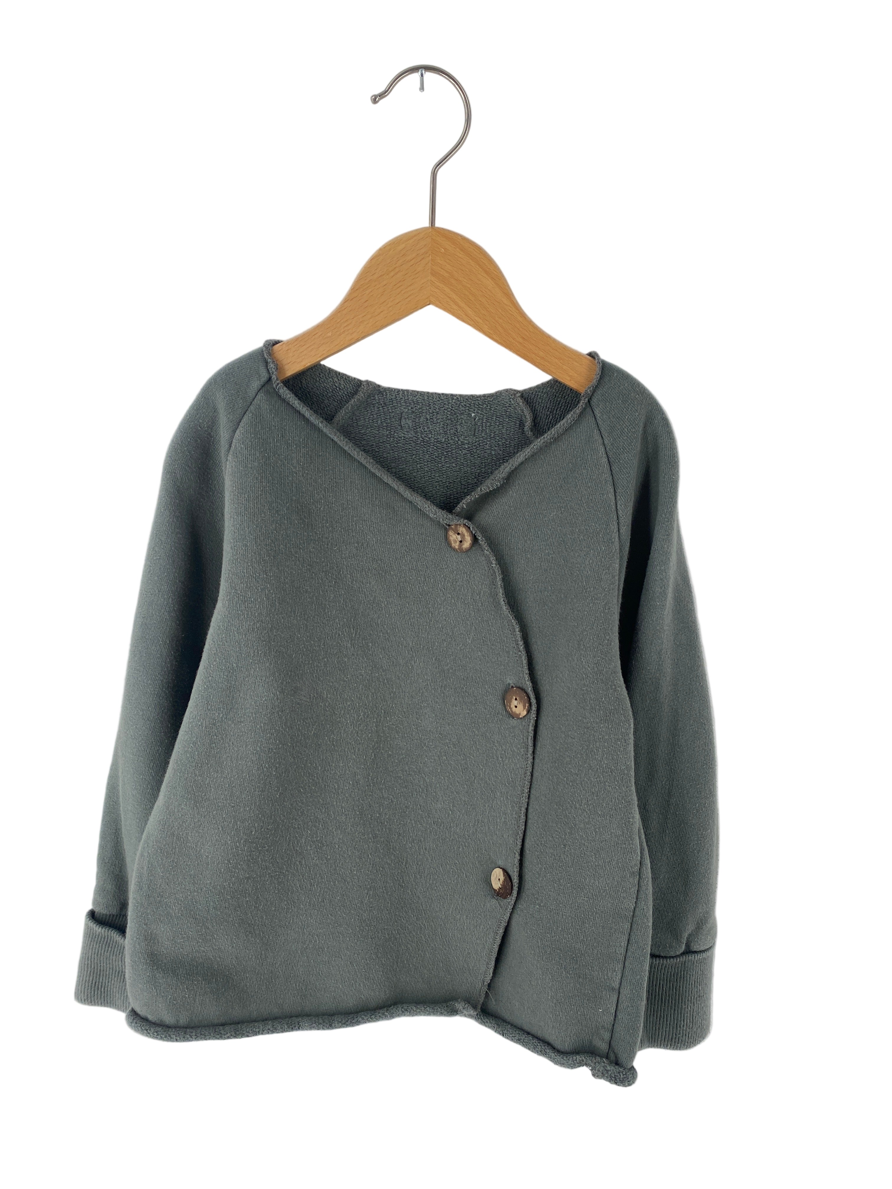 Schlüttli Bio-Sweat Taube Gr 3Y: Kleiner, gemütlicher cardigan in mutedem Grün mit dreifachen Holzknöpfen, langen Raglanärmeln, und rohen Kanten an Saum und Ausschnitt, ideal für Kleinkinder.