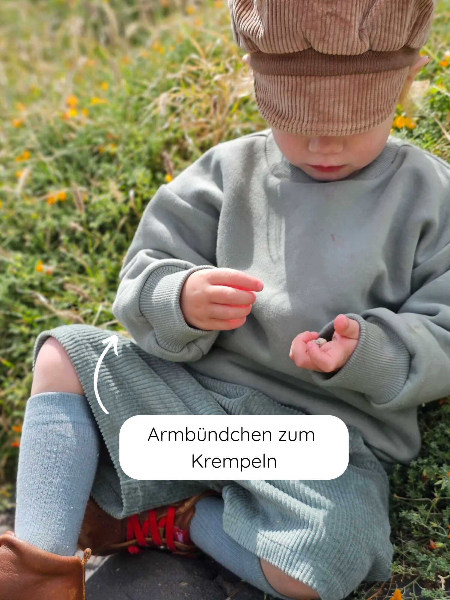 Kleinkind im grünen Pulli Bio-Sweat - Aqua mit aufgerollten Ärmeln, kniehohen Socken und braunen Stiefeln, spielt auf einer blumigen Wiese. Dezent und bequem für den Alltag.