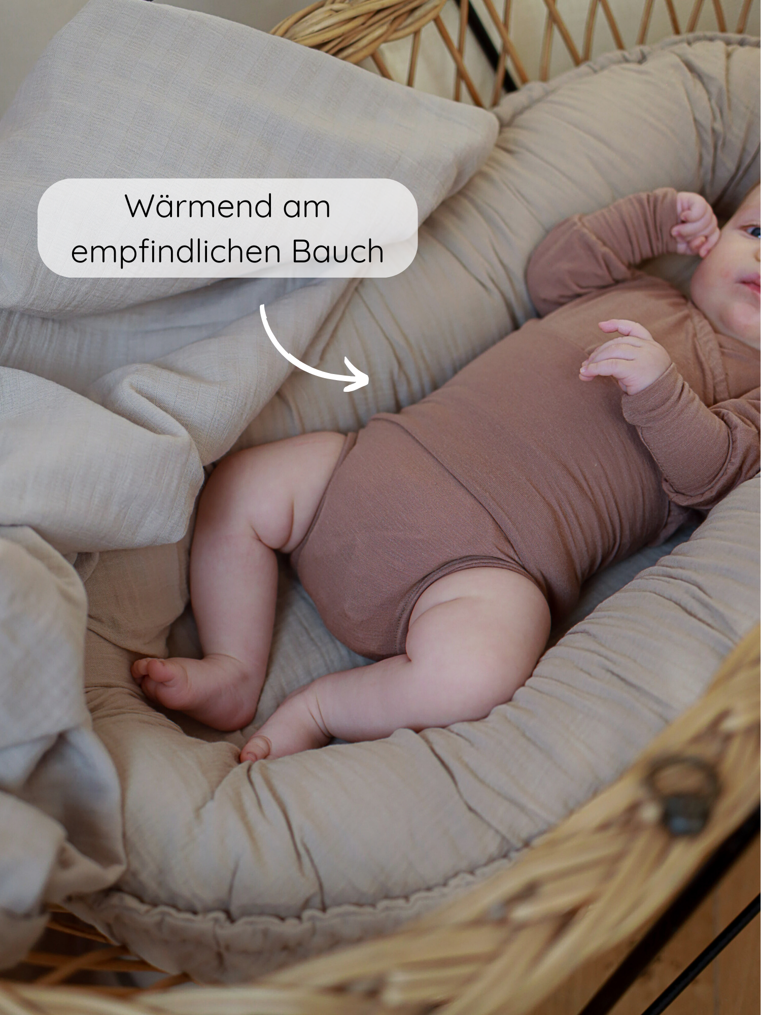 Ein Baby liegt in einem sanft gepolsterten Nest, trägt den Loopbody Kaschmir-Mix - Puder, langärmelig und ohne Knöpfe. Ein Text zeigt auf den wärmenden Effekt am Bauch.