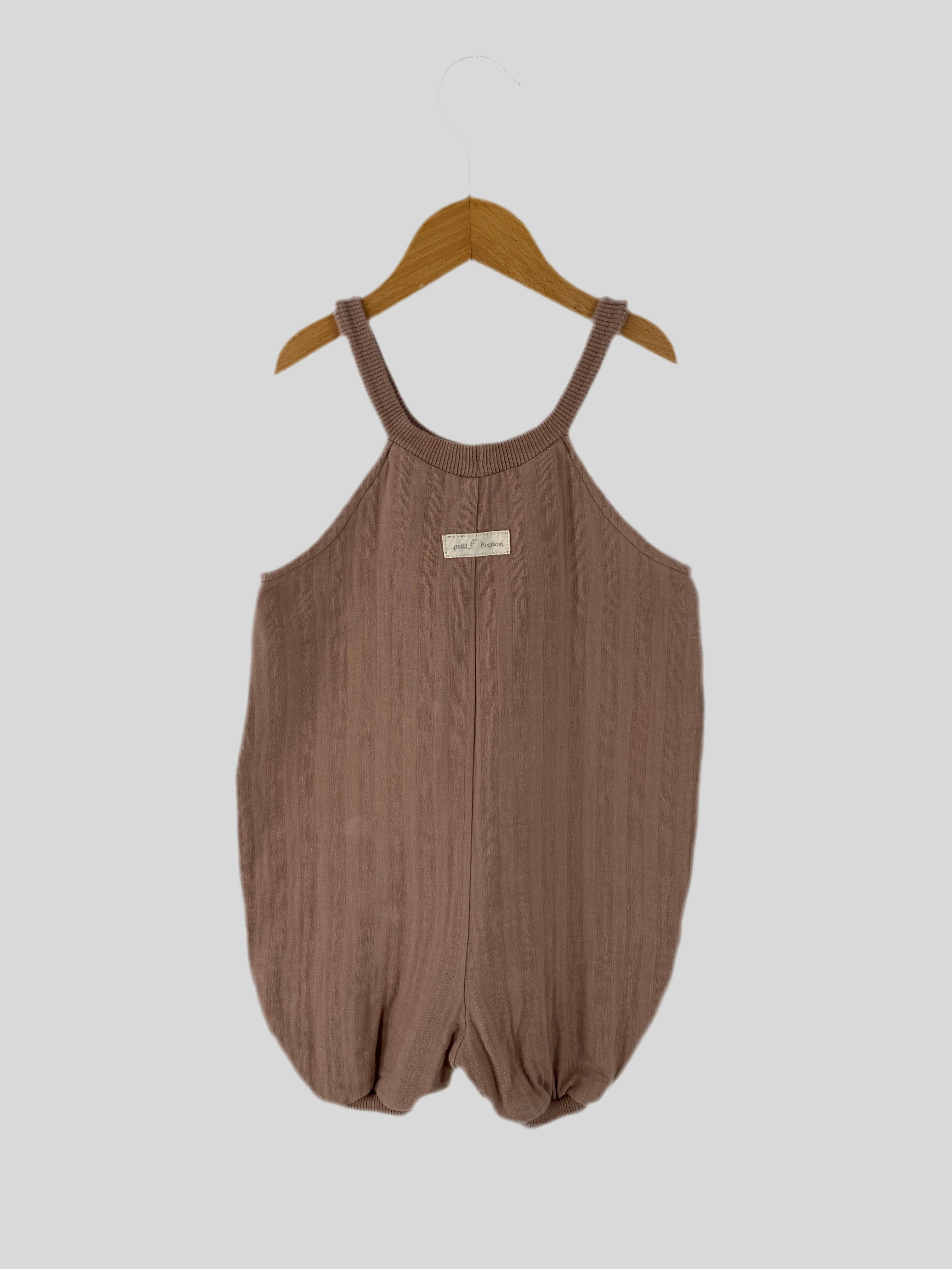 Kurze Schlupflatzhose Bio Musselin Puder Gr 1Y, ein ärmelloser, gestrickter Baby-Overall mit Rundhalsausschnitt und Bubbly-Beinöffnungen, hängt auf einem Holzbügel. Kleiner heller Fleck hinten sichtbar.