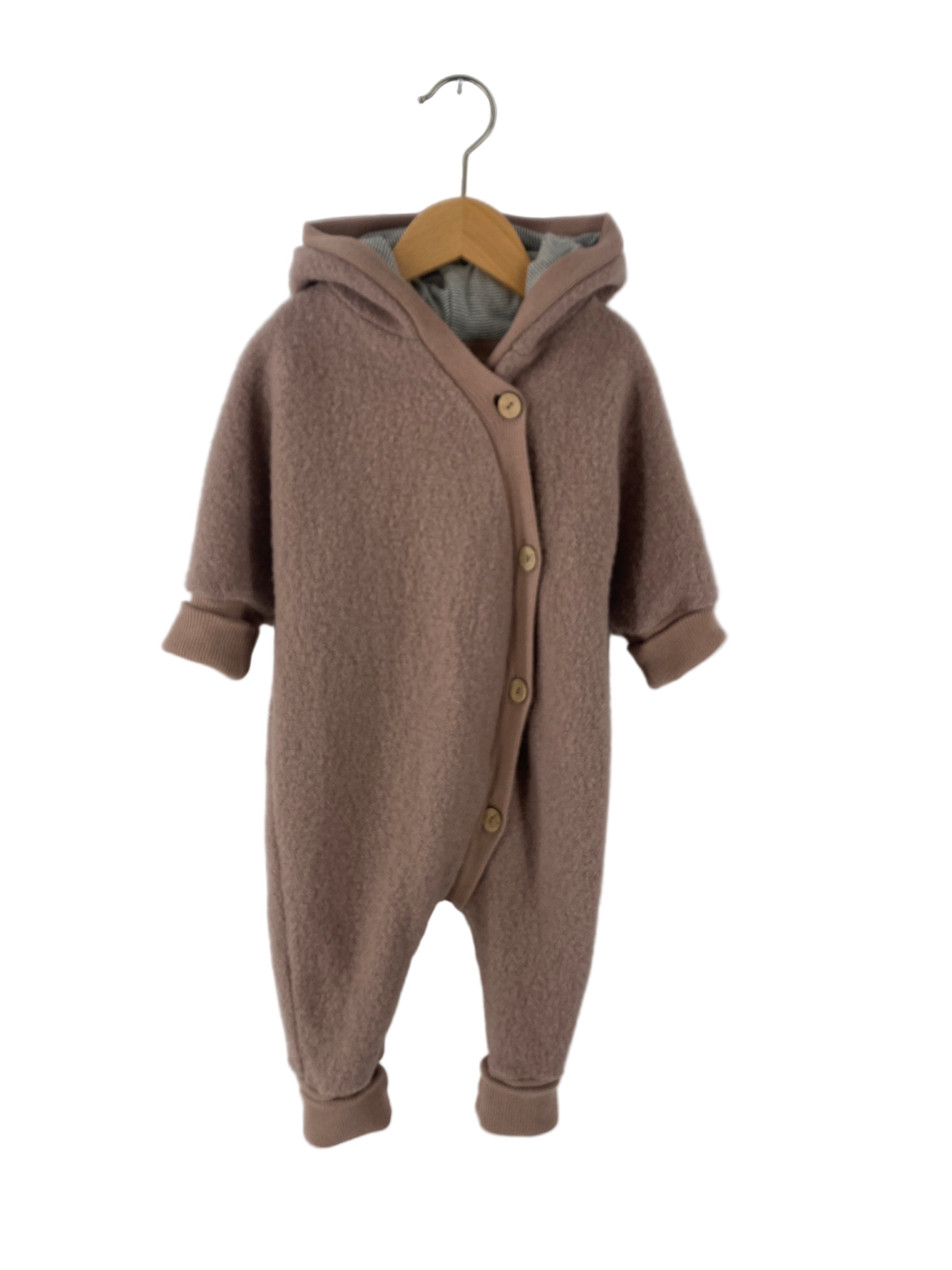 Kuschelanzug Wollwalk Puder Gr 6M: Ein weicher, einteiliger Baby-Overall mit Kapuze, langen Ärmeln, Bündchen und Knopfleiste, aus fleecigem Stoff, präsentiert auf einem Holzbügel vor weißem Hintergrund.