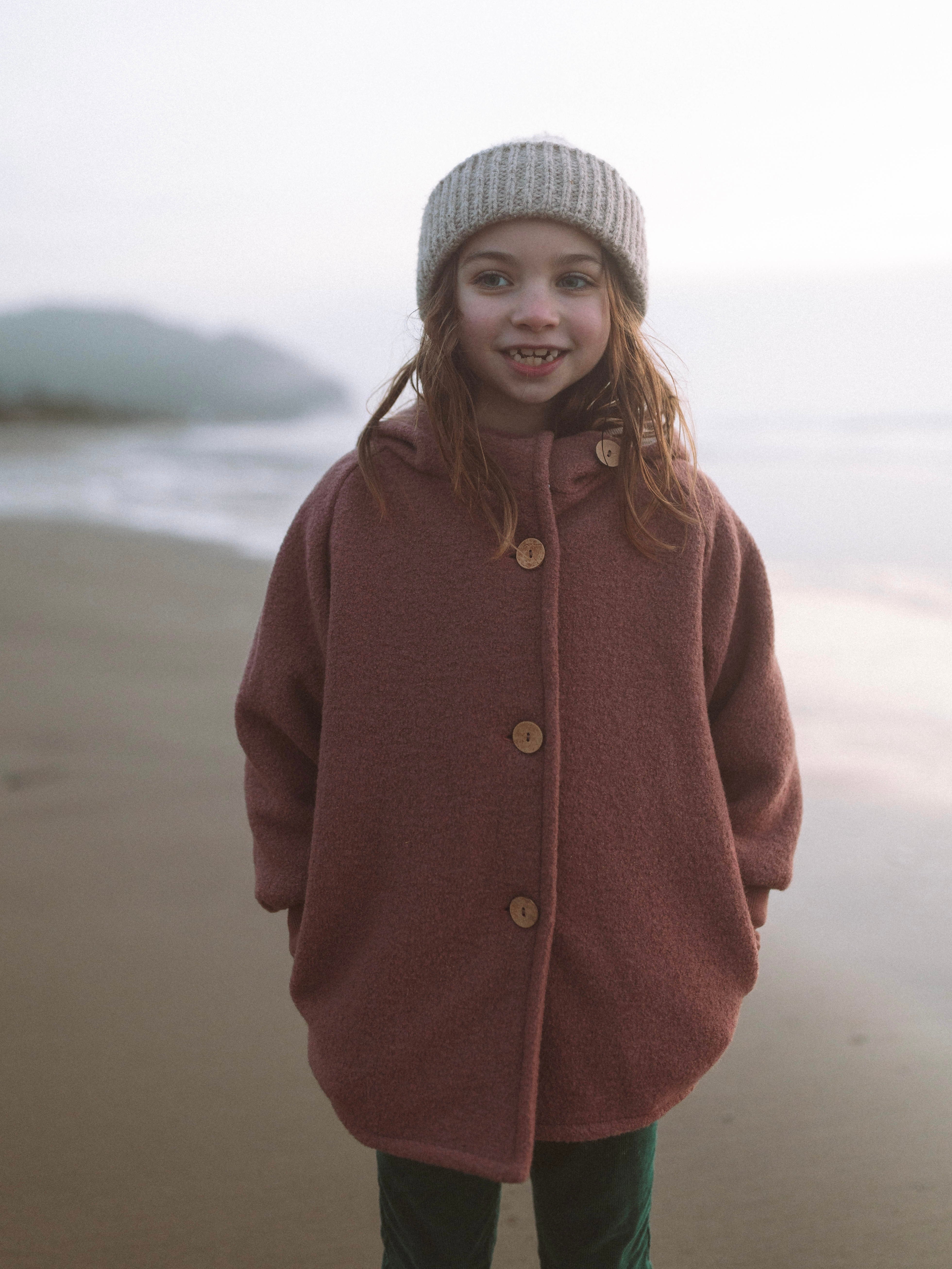 Ein Mädchen trägt die Wichteljacke Wollwalk - Zimt am Strand, mit hölzernen Knöpfen und Bio-Jersey-Futter, ergänzt durch eine graue Strickmütze, die Bewegungsfreiheit und Komfort bietet.