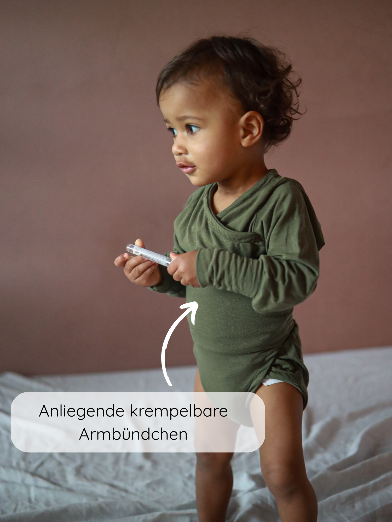 Ein Kleinkind trägt den Loopbody Kaschmir-Mix - Pistazie mit langen Ärmeln und anliegenden, krempelbaren Armbündchen, hält ein Handy und steht auf einem weichen Untergrund.