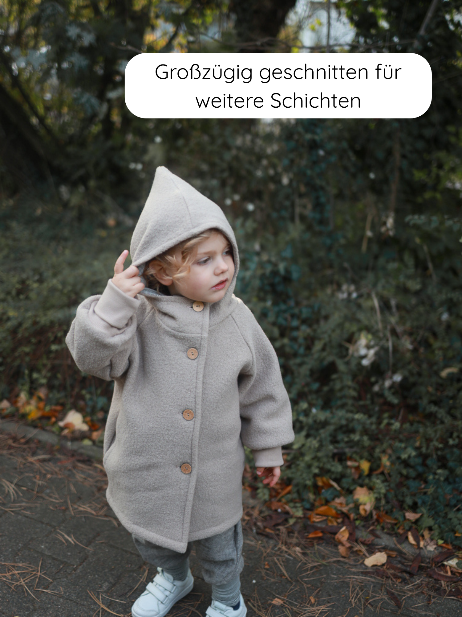 Kleinkind in der Wichteljacke Wollwalk - Sand mit Holzknöpfen, stehend auf einem Laubweg. Die großzügig geschnittene Jacke ermöglicht zusätzliche Schichten für kalte Tage.