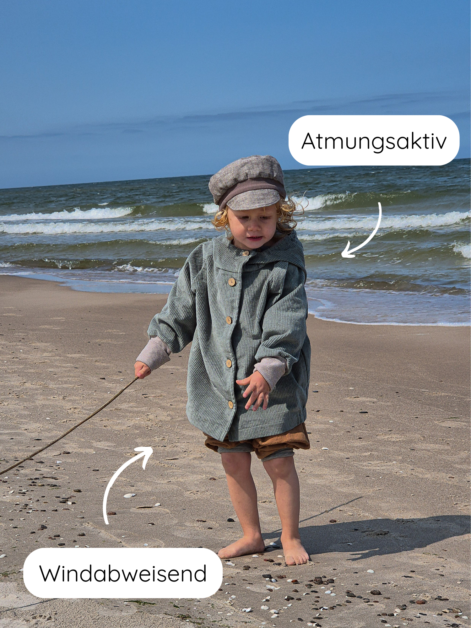 Kleinkind am Strand mit der Wichteljacke Breitcord Stretch- Aqua, atmungsaktiv und windabweisend, mit Holzknöpfen und passender Mütze, hält einen Stock, während es auf den Sand schaut.