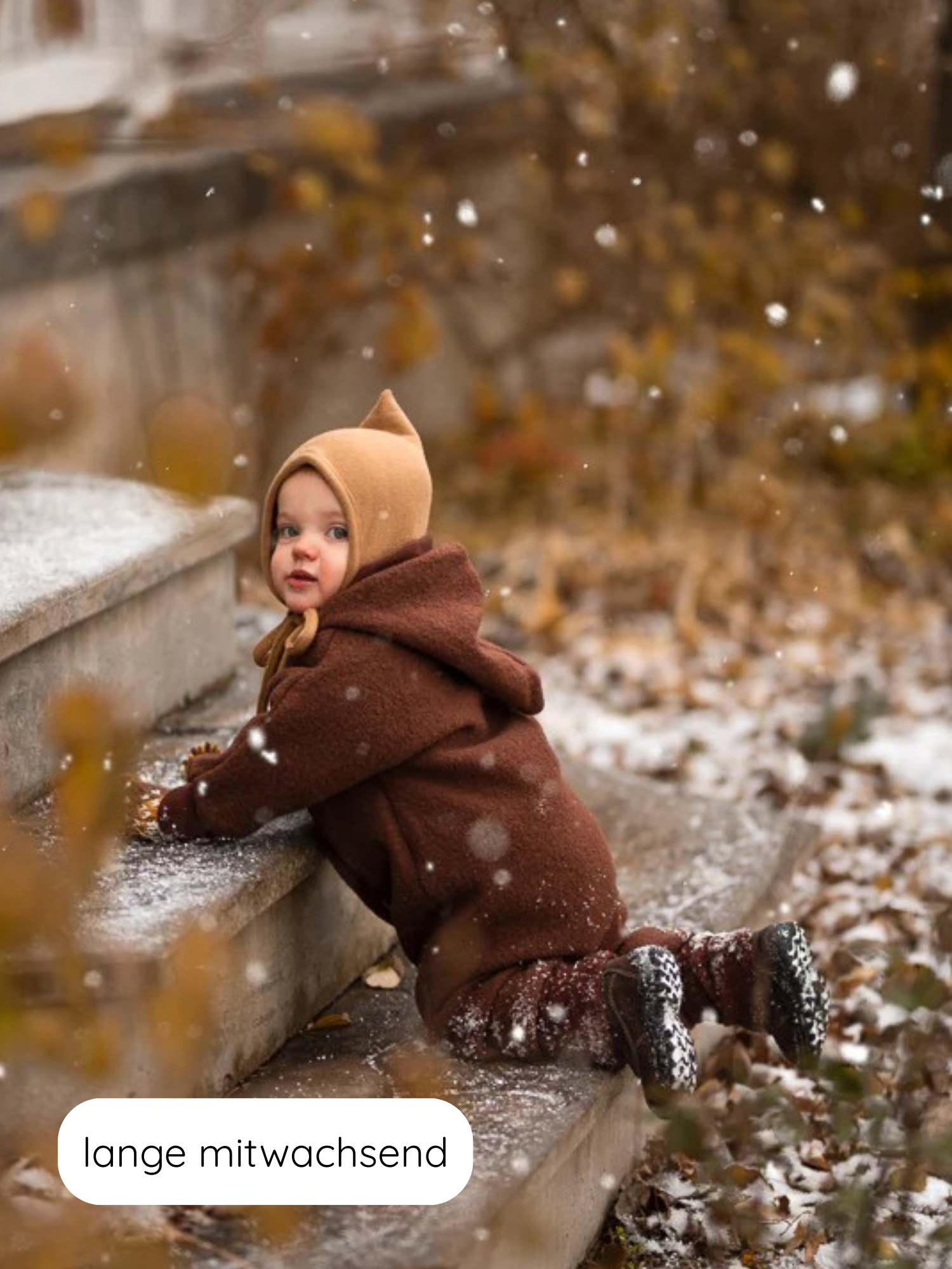 Kleinkind im Winter Wollwalk Overall mit Teddyfutter - Schoko, kniet auf schneebestäubtem Stein, umgeben von Laub. Der Overall bietet Wärme und Flexibilität durch anpassbare Bündchen für längere Nutzung.