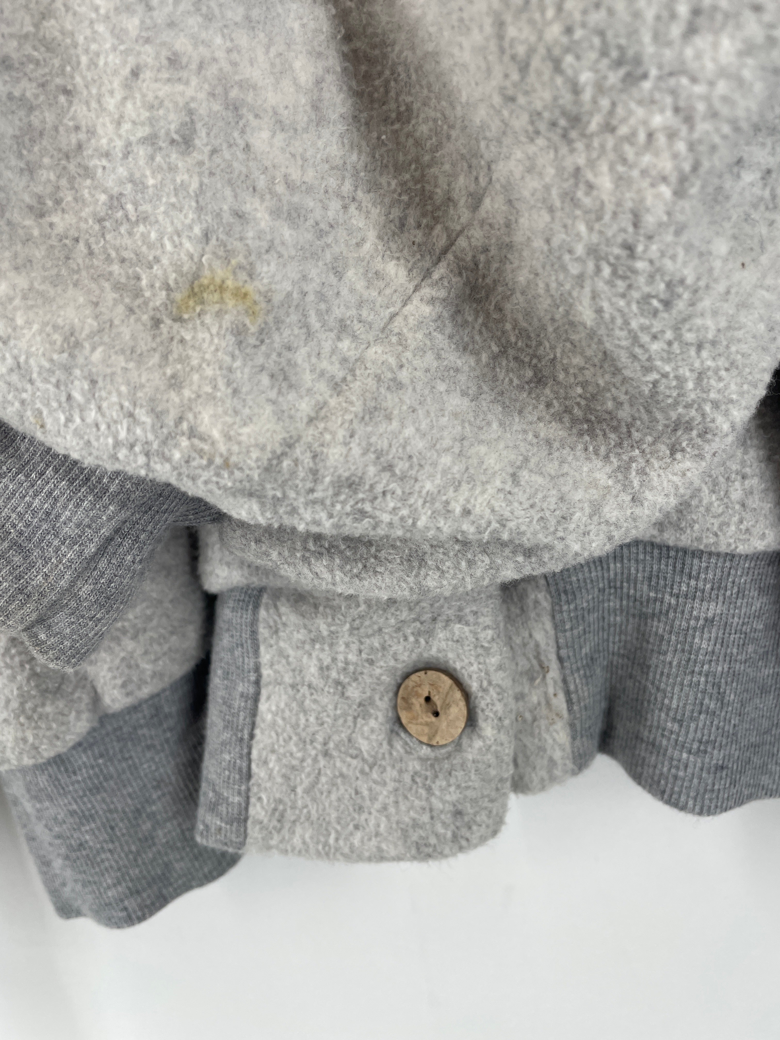 Nahaufnahme der Bomberjacke Plüsch Maus Gr. M zeigt einen hölzernen Zwei-Loch-Knopf am gerippten Strickbündchen mit einem kleinen gelblichen Fleck auf dem oberen Stoff. Zustand: gut, leichte Flecken.
