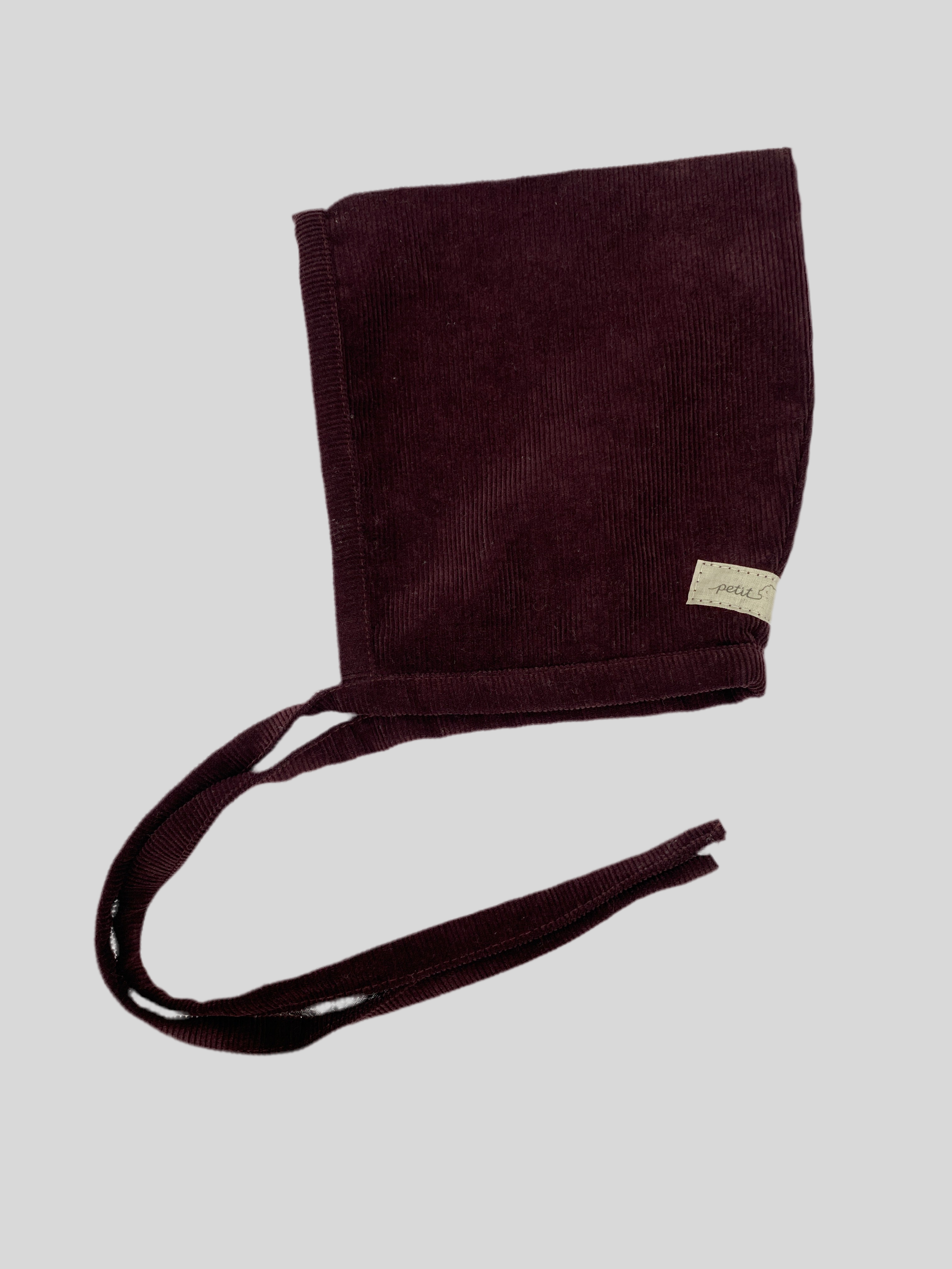 Zwergenmütze Feincord Aubergine Gr 6M: Kleines, ungefüttertes Cord-Babyhäubchen mit langen Stoffbändern zum Binden und einem „petit“-Label seitlich.