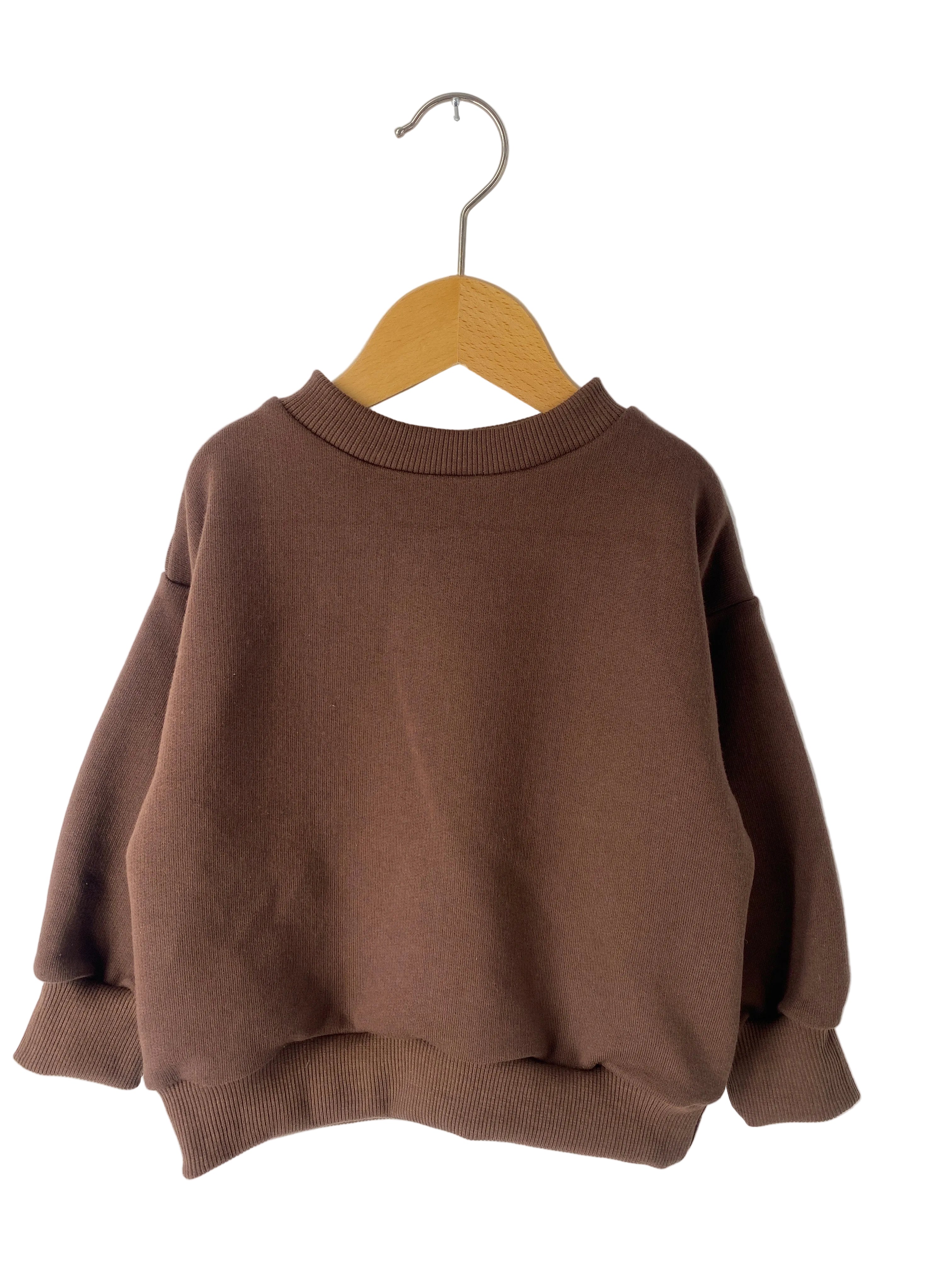 Der Pulli Bio Sweat Schoko Gr 1Y, ein neuer brauner Pullover, hängt an einem Holzkleiderbügel. Ein leichter Strich ist im Stoff vorne sichtbar.
