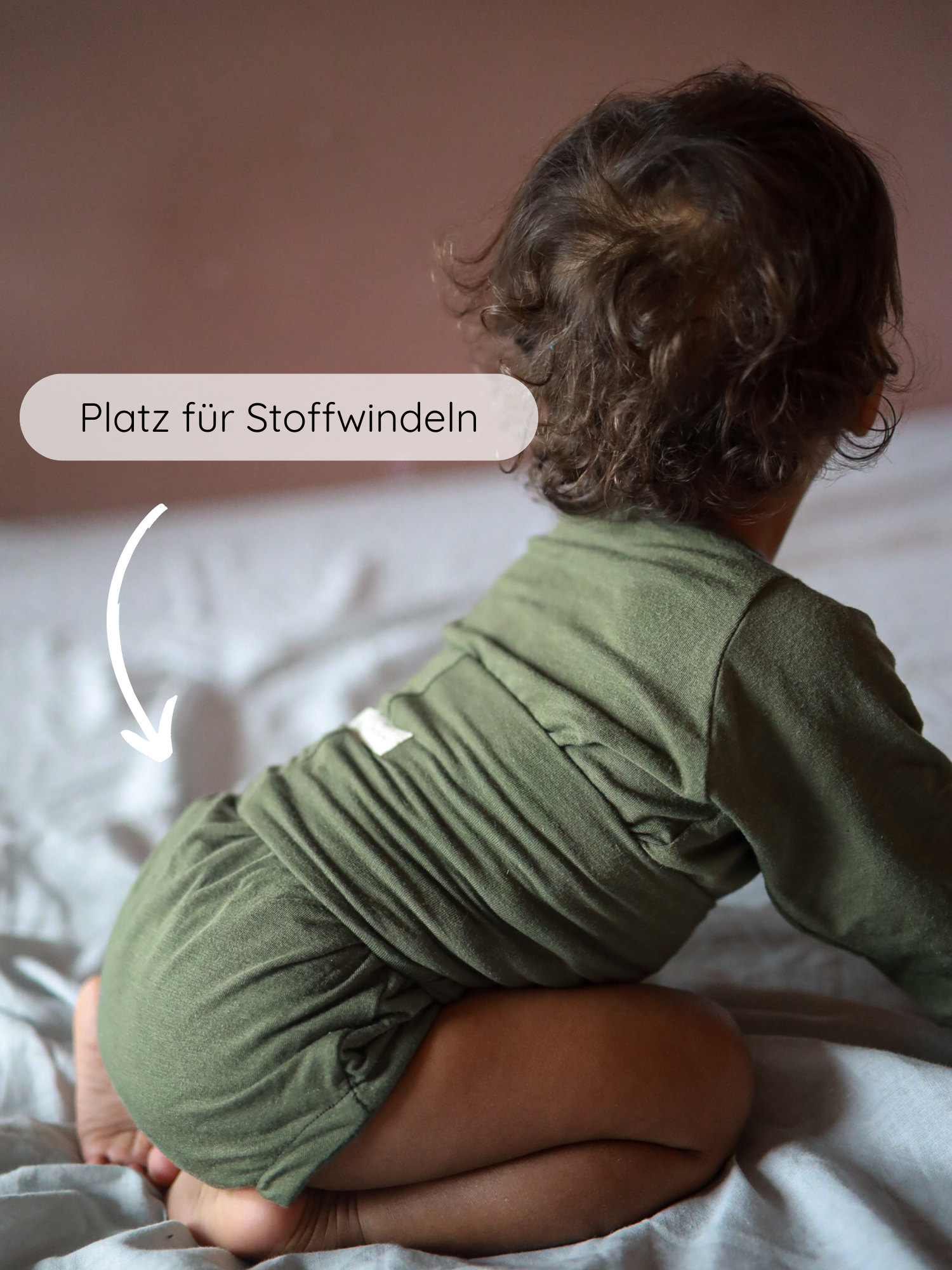 Kleinkind trägt Loopbody Kaschmir-Mix - Pistazie, kniet auf weicher Oberfläche. Pfeil zeigt auf Platz für Stoffwindeln im Rückenbereich, unterstreicht extra Raum für bequeme Passform mit Stoffwindeln.