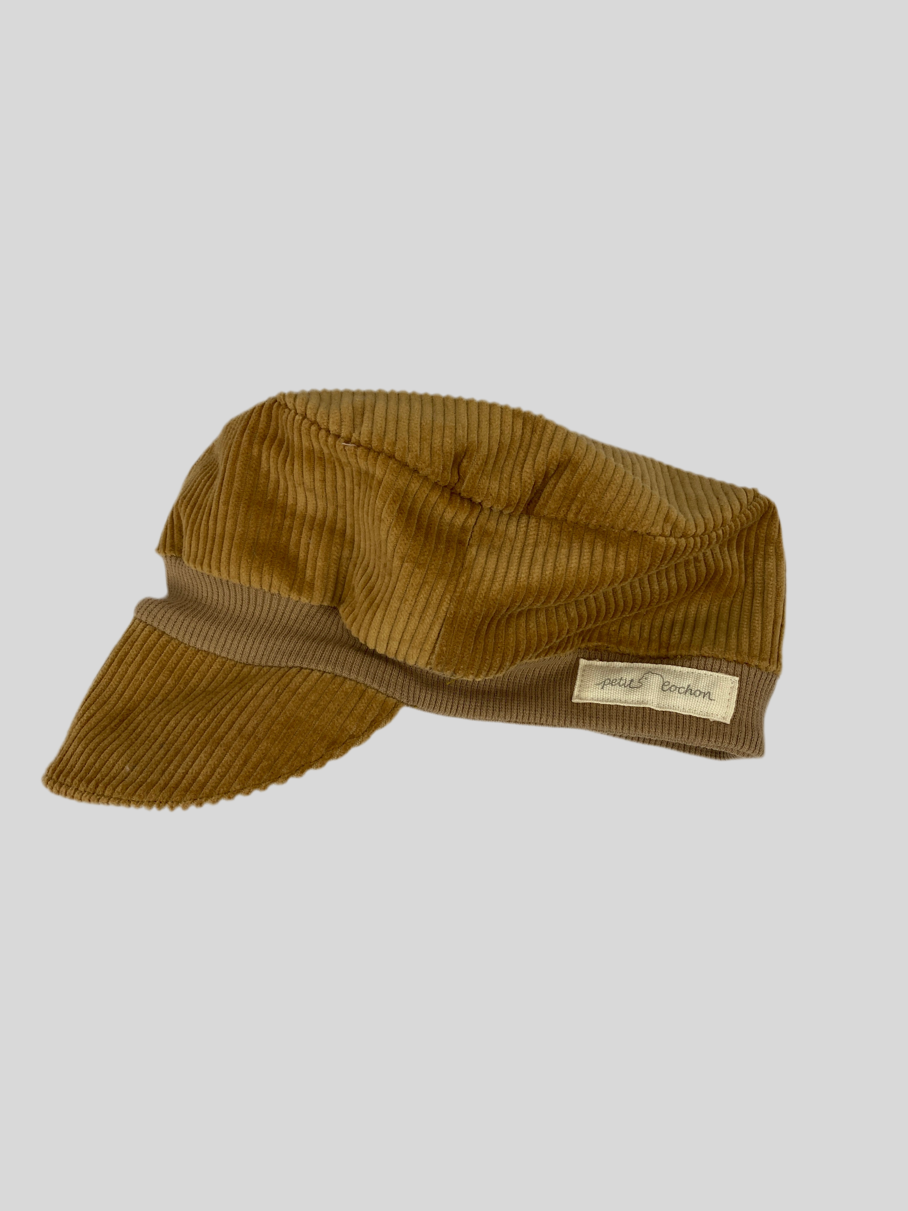 Kopie Michelmütze Breitcord beige Gr 6M-2Y, eine braune Cord-Mütze im Militärstil mit kurzem, gebogenem Schirm und kleinem „petit cochon“-Etikett am Band, vor hellem grauen Hintergrund.