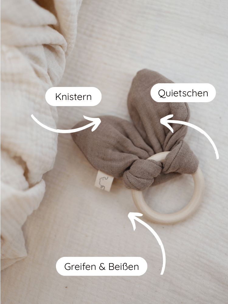 Baby Beißring Baumwoll-Musselin - Praline, zeigt einen glatten Holzring mit knisternden Musselin-Knoten. Integrierter Quietscher, ideal zum Greifen und Beißen, fördert sensorische Entwicklung. Nachhaltig, speichelfest, handgefertigt in Berlin.