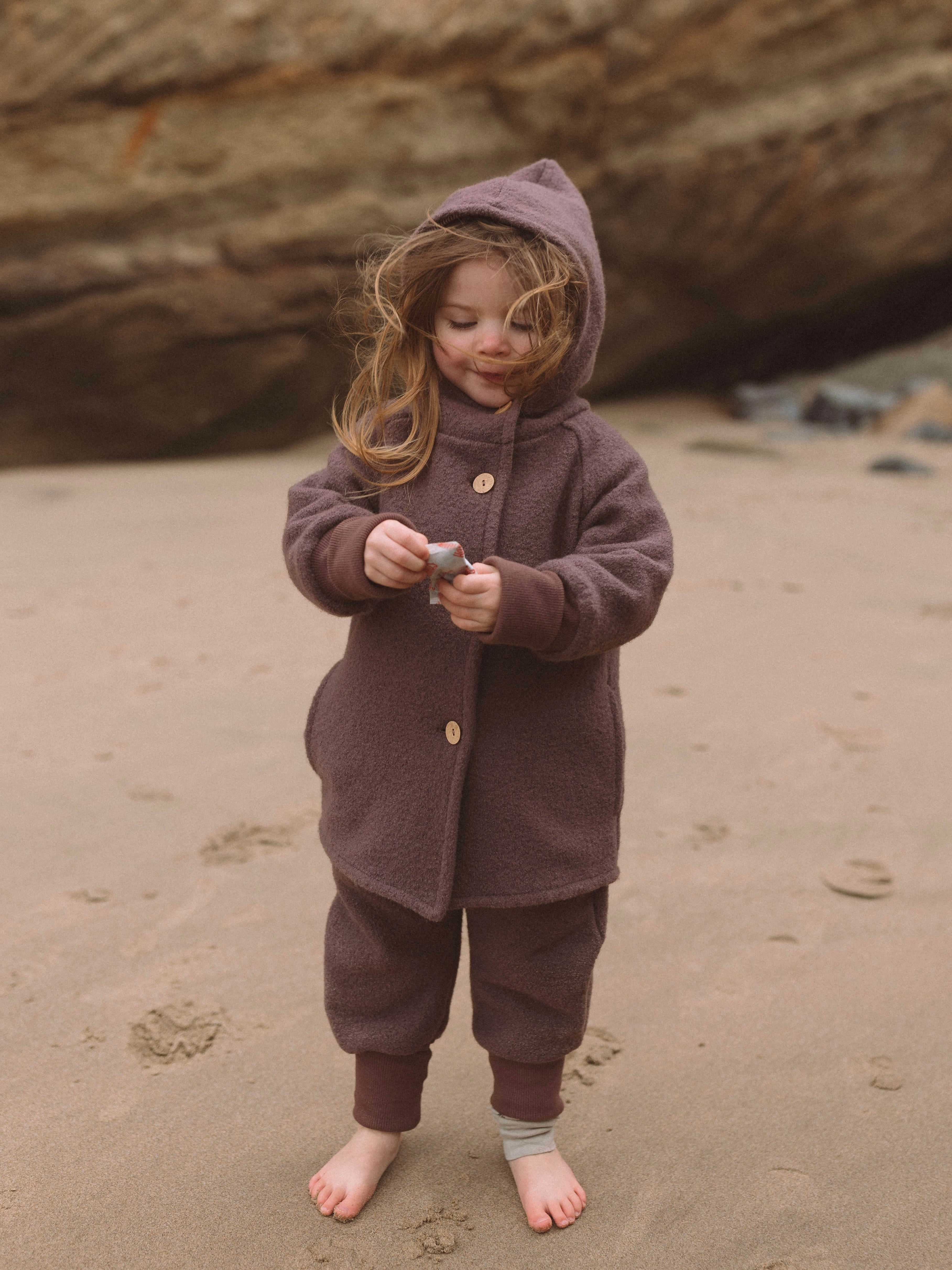 Kleinkind am Strand, trägt die Wichteljacke Wollwalk - Malve, hält ein kleines Objekt. Die Jacke bietet Wärme, Bewegungsfreiheit und ist ideal für Herbst, Winter und Frühling.