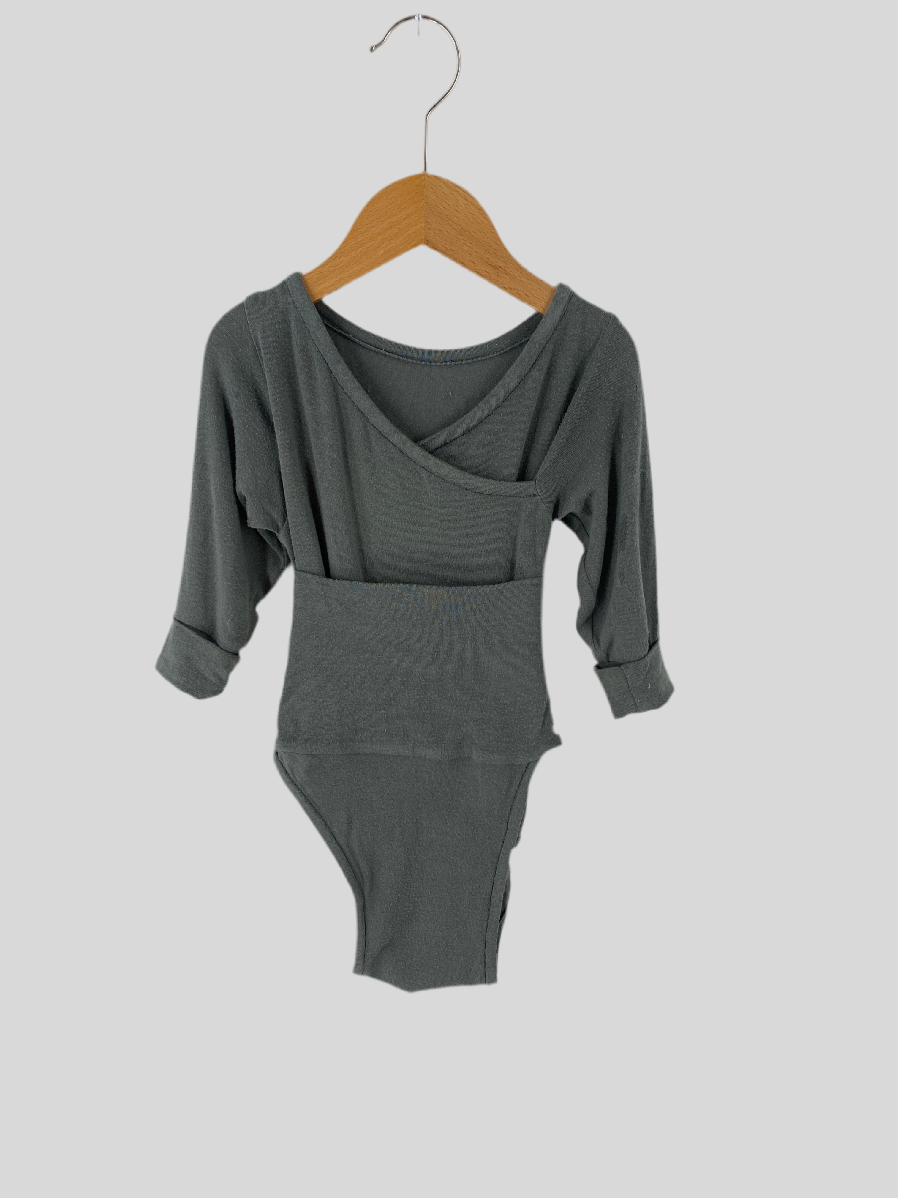 Loopbody Kaschmir Mix Taube Gr 3M: Ein graugrüner Langarm-Body mit weitem Taillenbund, an einem Holzkleiderbügel hängend. Zustand: Gut, leichte Gebrauchsspuren mit 2-3 kleinen Löchern im Stoff.