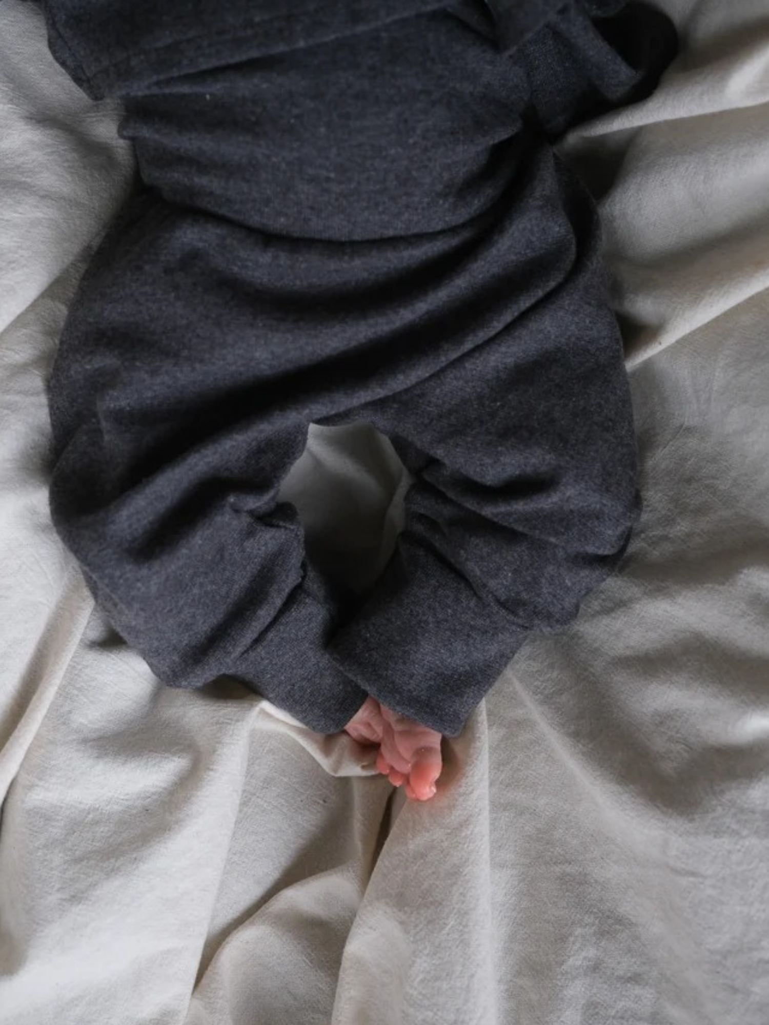 Babyknicker Feinstrick - Ruß Meliert, zeigt die unteren Beine eines Babys, das auf einer weichen Oberfläche liegt, mit dunklen, leicht lockeren Hosen, aus denen kleine, nackte Füße hervorschauen.
