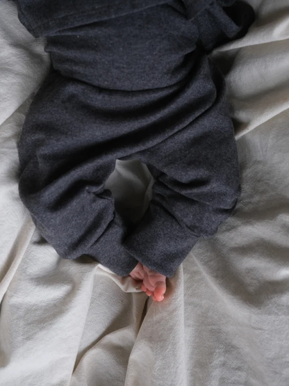Babyknicker Feinstrick - Ruß Meliert, zeigt die unteren Beine eines Babys, das auf einer weichen Oberfläche liegt, mit dunklen, leicht lockeren Hosen, aus denen kleine, nackte Füße hervorschauen.