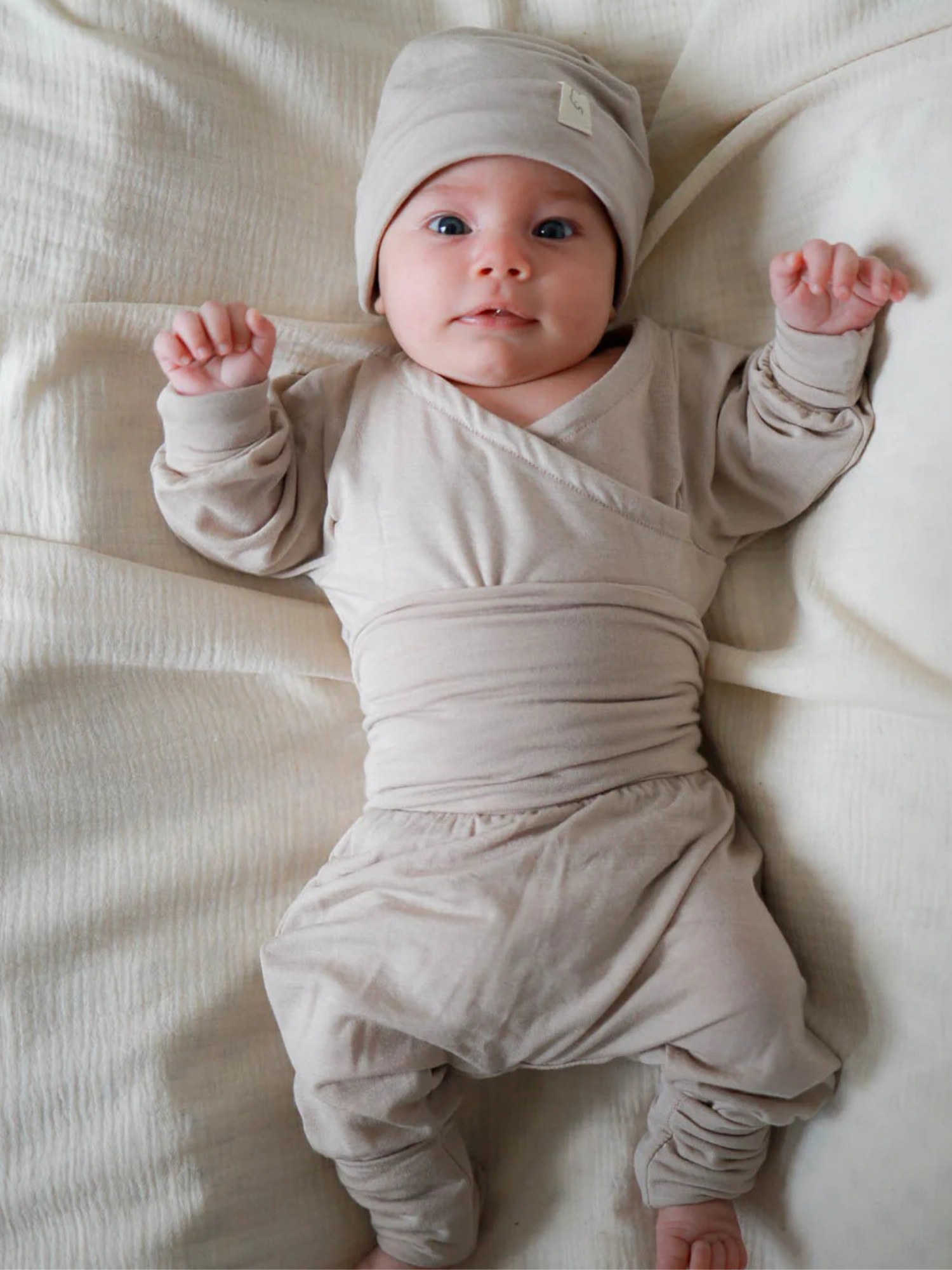 Baby in beigefarbener Babyknicker Kaschmir-Mix - Sand mit passender Mütze, auf weicher Unterlage liegend, Arme leicht angehoben, zeigt die weiche, wärmende und atmungsaktive Qualität der Kleidung.