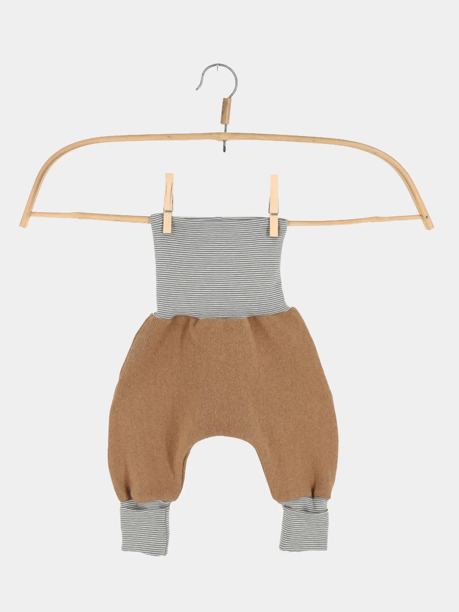 Babyknicker Strick - Sahara mit Ringelbündchen, hängend an einem Holzkleiderbügel mit Clips. Die Hose hat gestreifte Bündchen und einen warmen, camelbraunen Strickkörper.