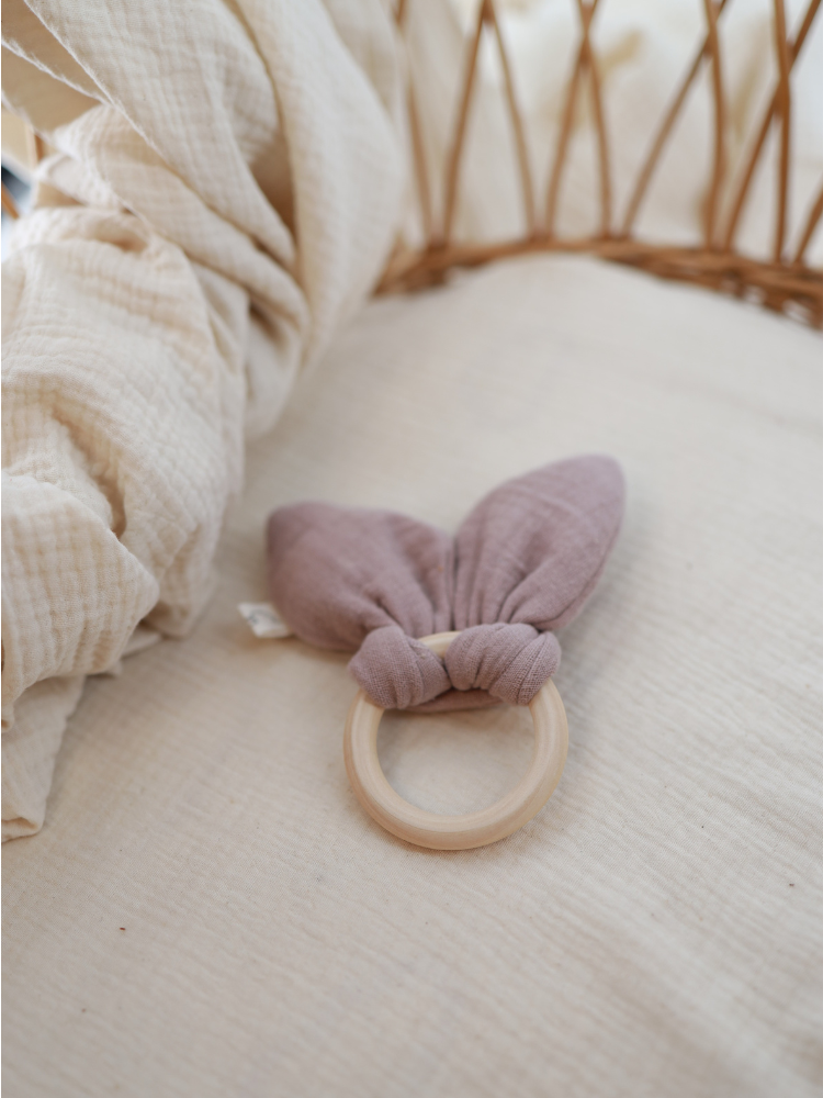 Baby Beißring Baumwoll-Musselin - Puder: Ein glatter Holzring mit angeknotetem, weichem Musselin-Bunny Ear-Tuch, ideal als Zahnungshilfe und Fühlspielzeug für Babys, auf hellem Stoff präsentiert.