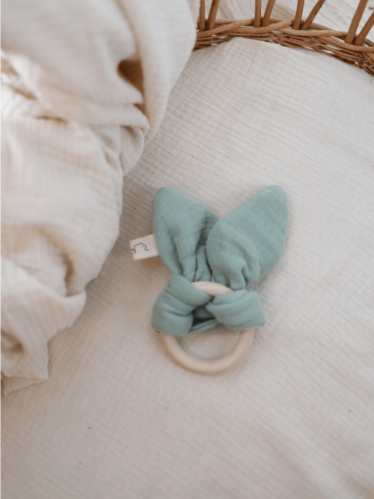 Baby Beißring Baumwoll-Musselin - Mint liegt auf einem cremefarbenen Untergrund, bestehend aus einem glatten Holzring und weichen, geknoteten Musselin-Ohren, ideal für zahnende Babys.