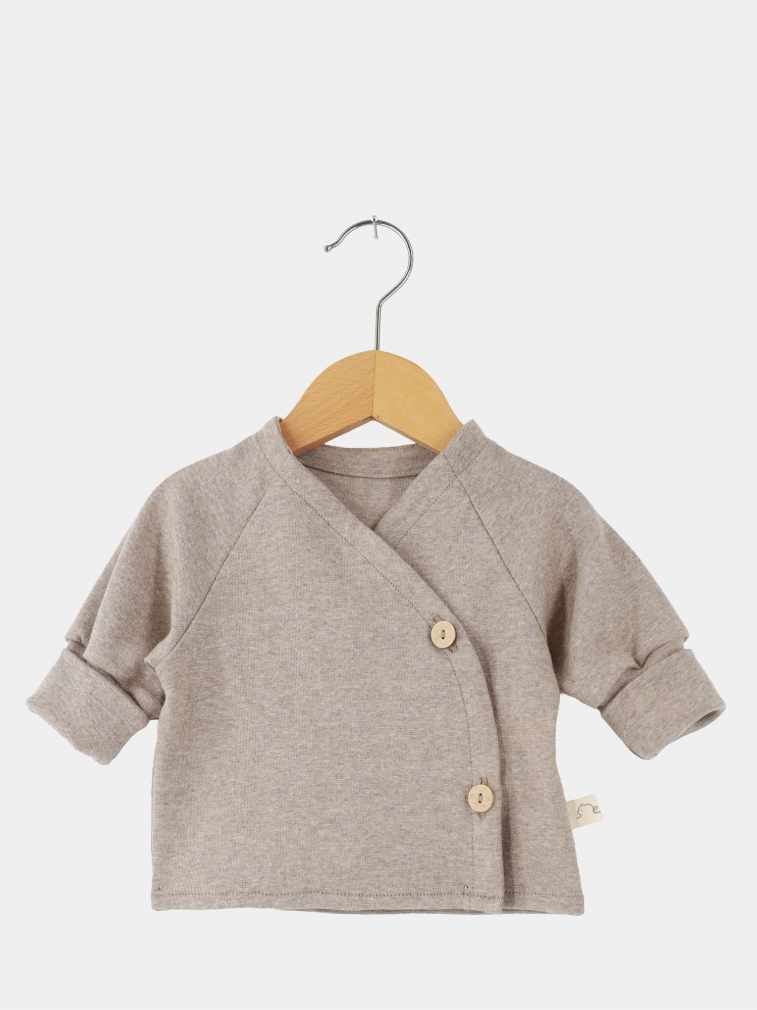 Babyschlüttli Feinstrick - Hafer Meliert: Ein Wickel-Baby-Strickpullover in neutralem Beige, mit langen Ärmeln und Holzknöpfen auf einer Seite, hängt auf einem Holzbügel. Ideal für zarte Babyhaut.