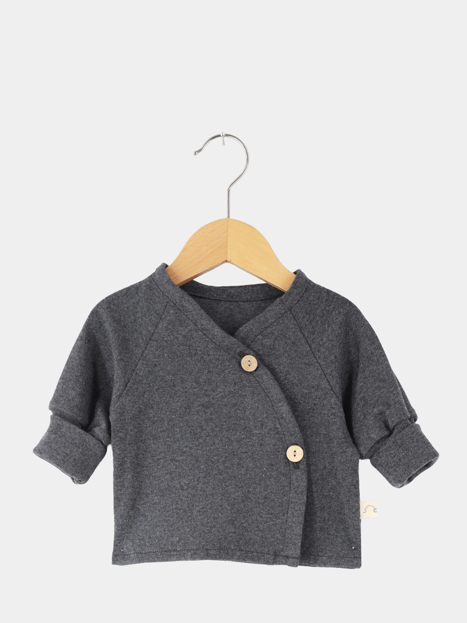 Babyschlüttli Feinstrick - Ruß Meliert: Dunkelgraue Strickjacke mit asymmetrischem Wickelverschluss, zwei Holzknöpfen und hochgekrempelten Ärmeln, präsentiert auf einem Holzbügel vor hellem Hintergrund.