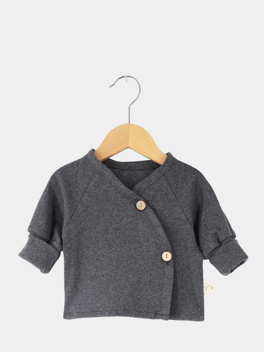 Babyschlüttli Feinstrick - Ruß Meliert: Dunkelgraue Strickjacke mit asymmetrischem Wickelverschluss, zwei Holzknöpfen und hochgekrempelten Ärmeln, präsentiert auf einem Holzbügel vor hellem Hintergrund.