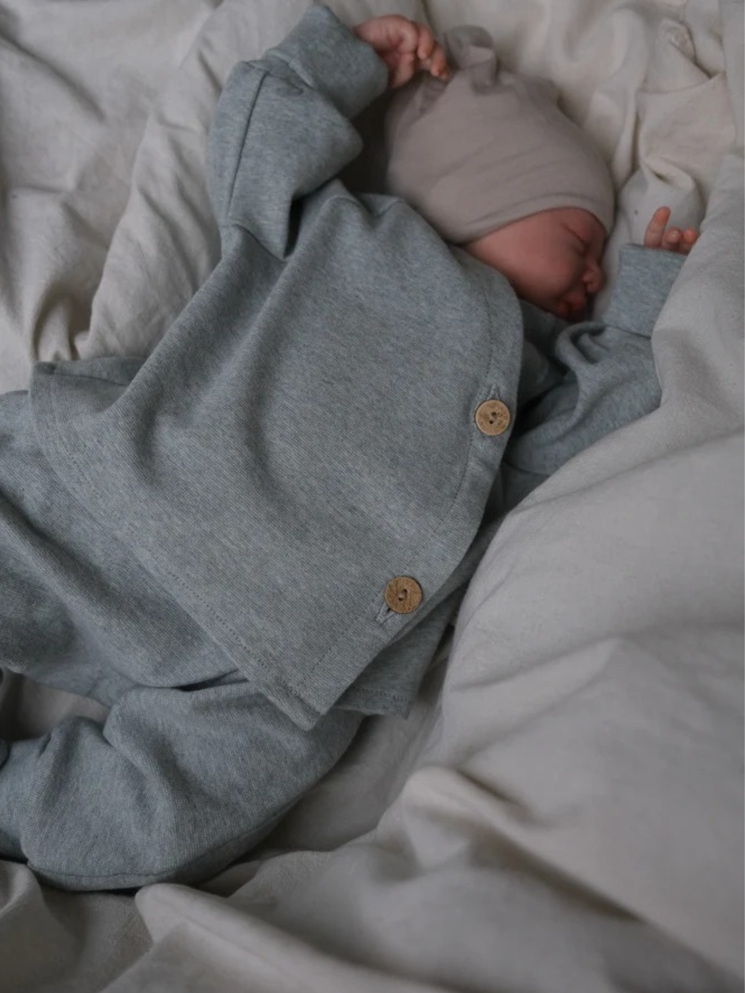 Ein Neugeborenes schläft in einem grauen Babyschlüttli Feinstrick - Mint Meliert, mit zwei Holzknöpfen, auf weicher, neutraler Bettwäsche.