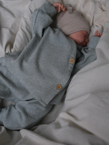 Ein Neugeborenes schläft in einem grauen Babyschlüttli Feinstrick - Mint Meliert, mit zwei Holzknöpfen, auf weicher, neutraler Bettwäsche.